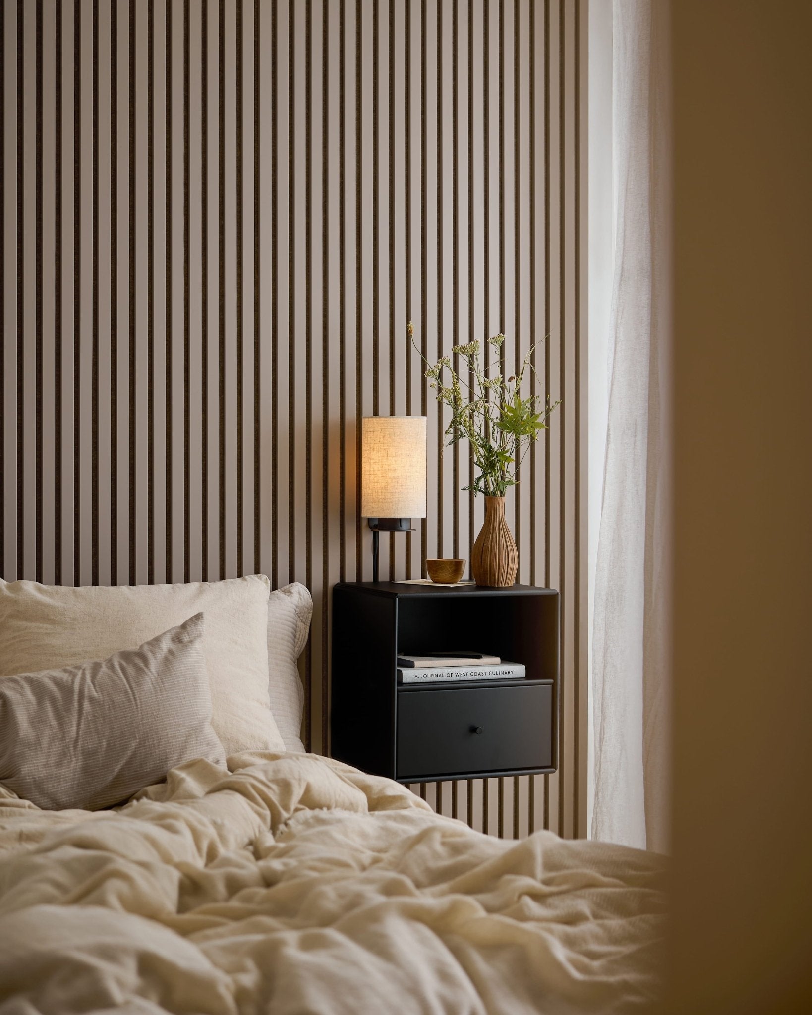 Una cama con ropa de cama color crema junto a una mesita de noche negra, delante de una pared de Akupanel en Dark Cashmere.