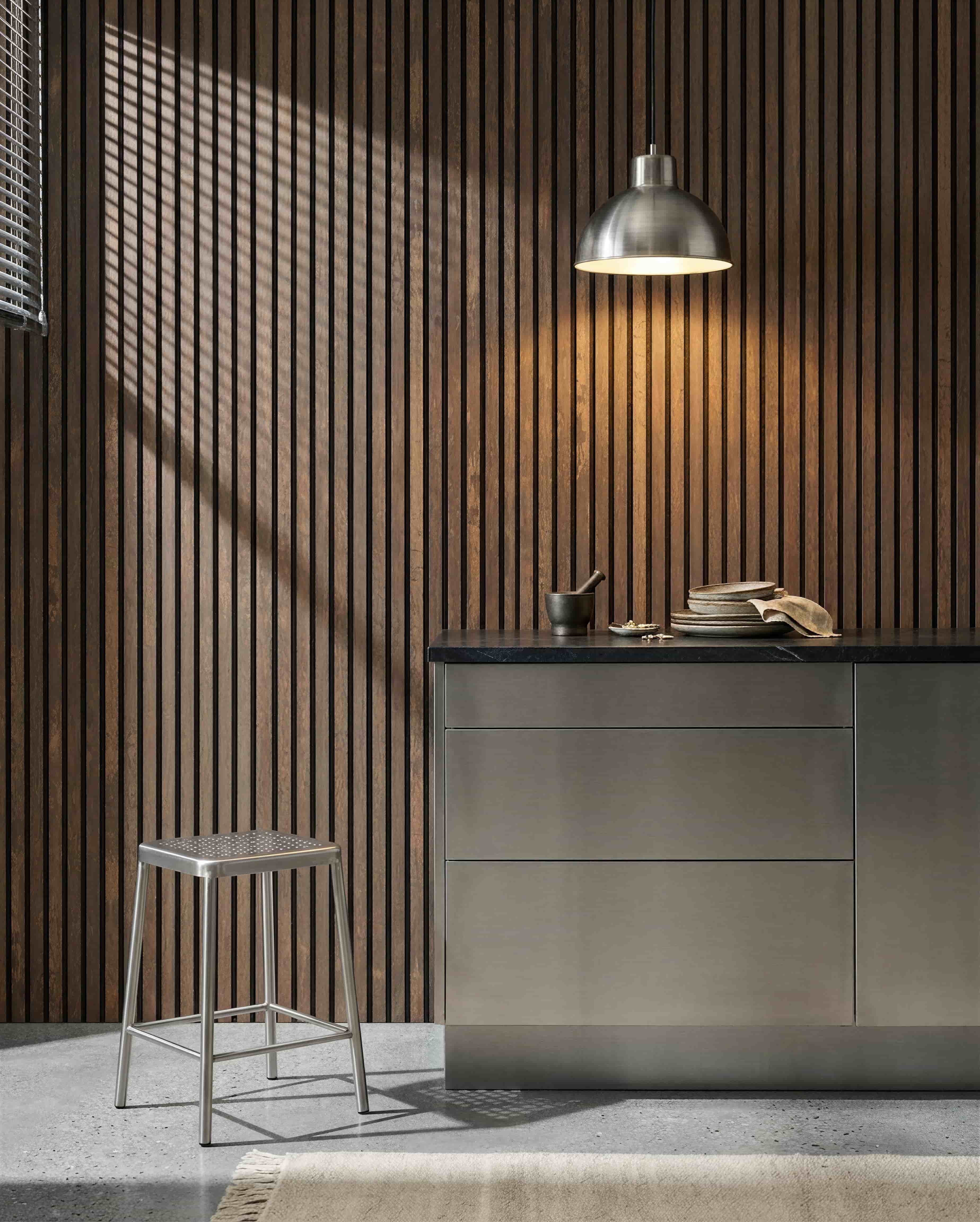 Cocina con una pared acústica de listones WoodUpp Akupanel Oxide Copper, encimera de acero inoxidable, taburete de metal, lámpara colgante, vajilla variada y mortero.