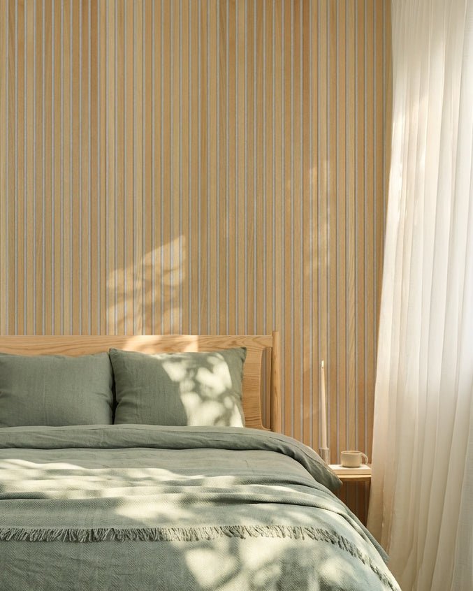 Dormitorio nórdico minimalista con ropa de cama verde salvia y un panel acústico de lamas WoodUpp Classic Oak Akupanel; la luz del sol crea sombras suaves sobre la cama.