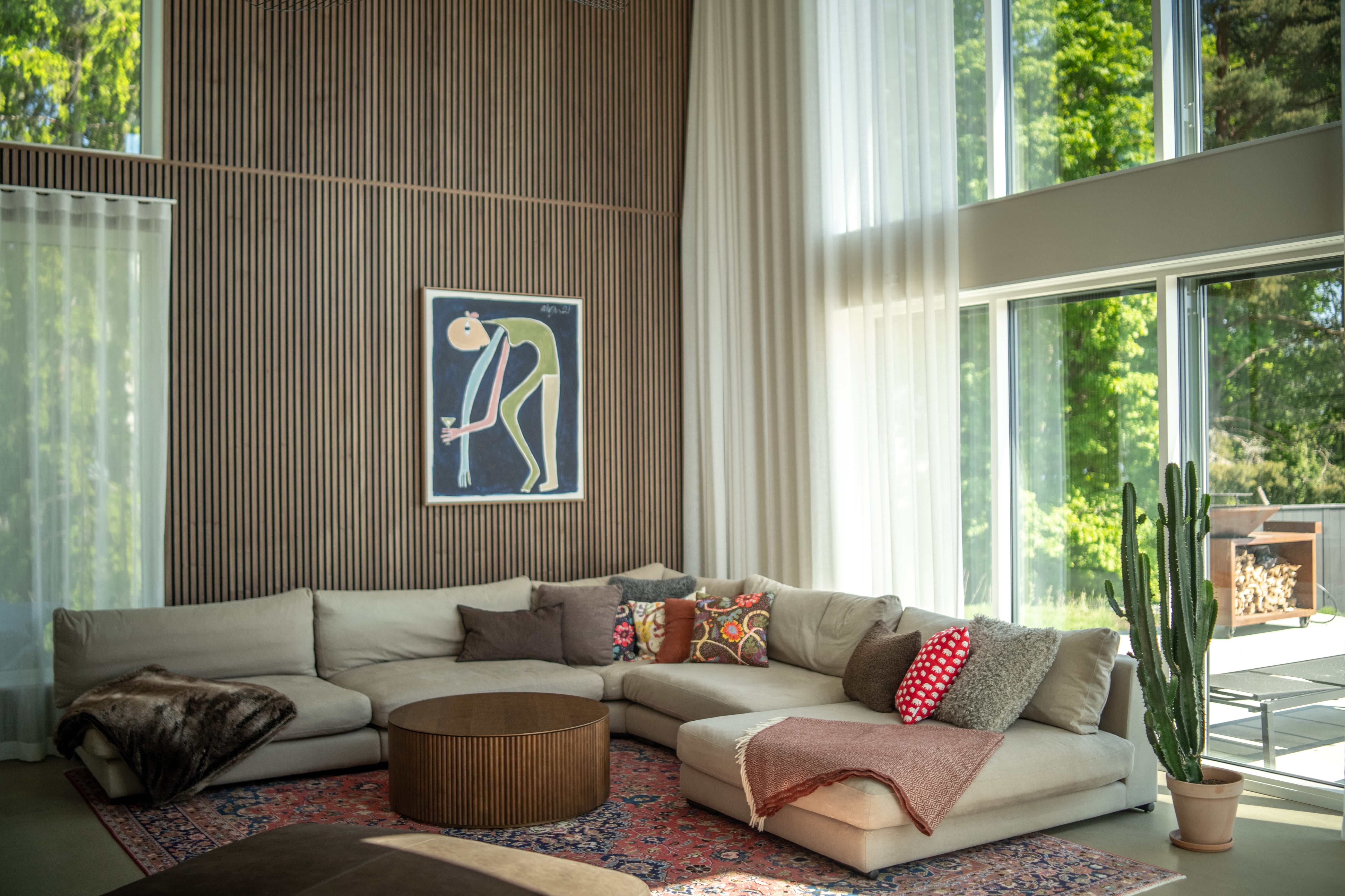 Moderno salón con un gran sofá seccional beige, cojines de colores, mesa de centro redonda, cactus y ventanas del suelo al techo con cortinas transparentes y paneles acústicos nórdicos.
