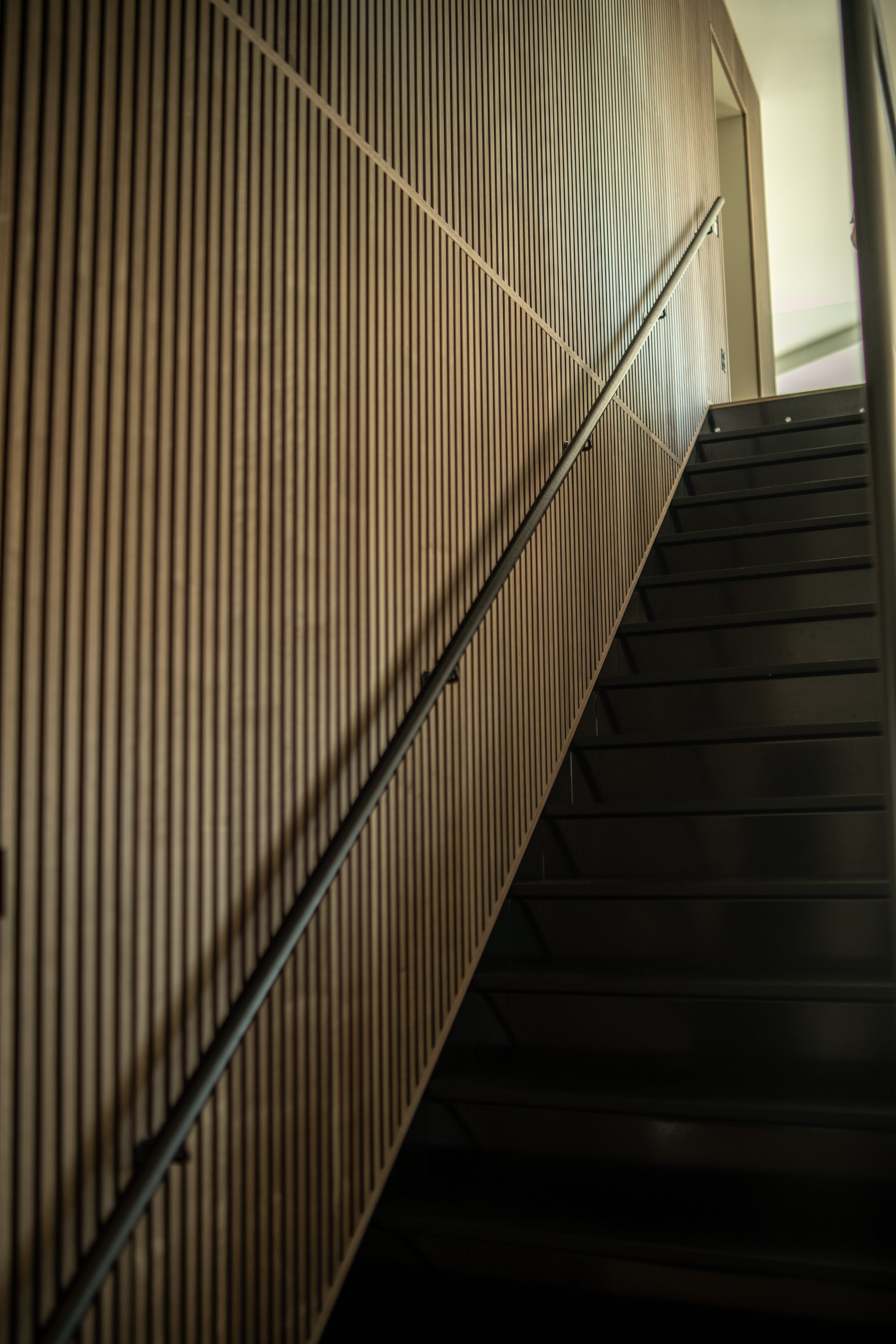 Una escalera moderna con peldaños negros se complementa con un panel vertical de lamas acústicas, visto desde abajo en ángulo para conseguir un aspecto nórdico minimalista.