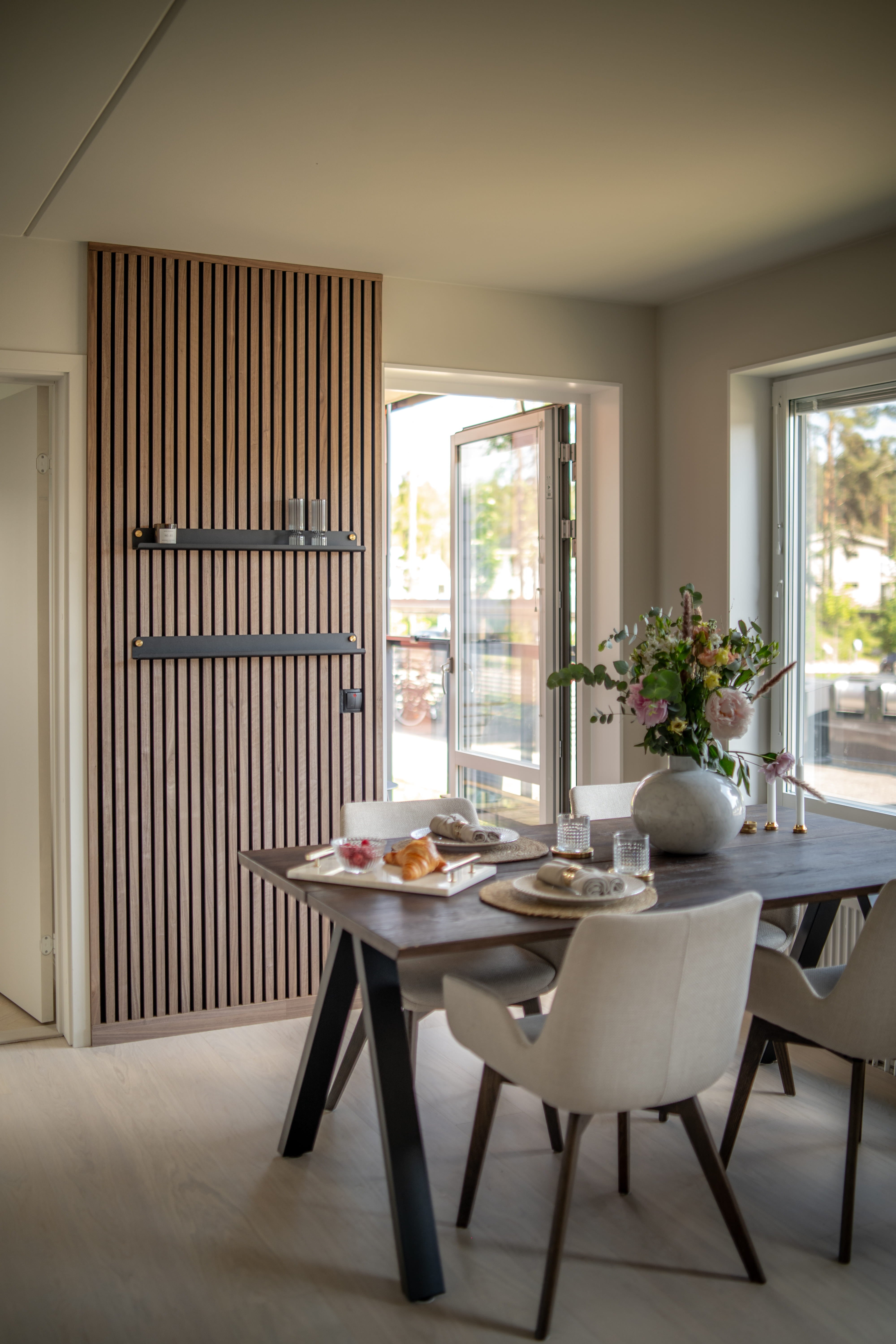 Comedor moderno con pared de listones de madera acústica Akupanel texturizados, mesa de desayuno oscura, sillas minimalistas de color beige y un jarrón de flores junto a grandes ventanales abiertos.