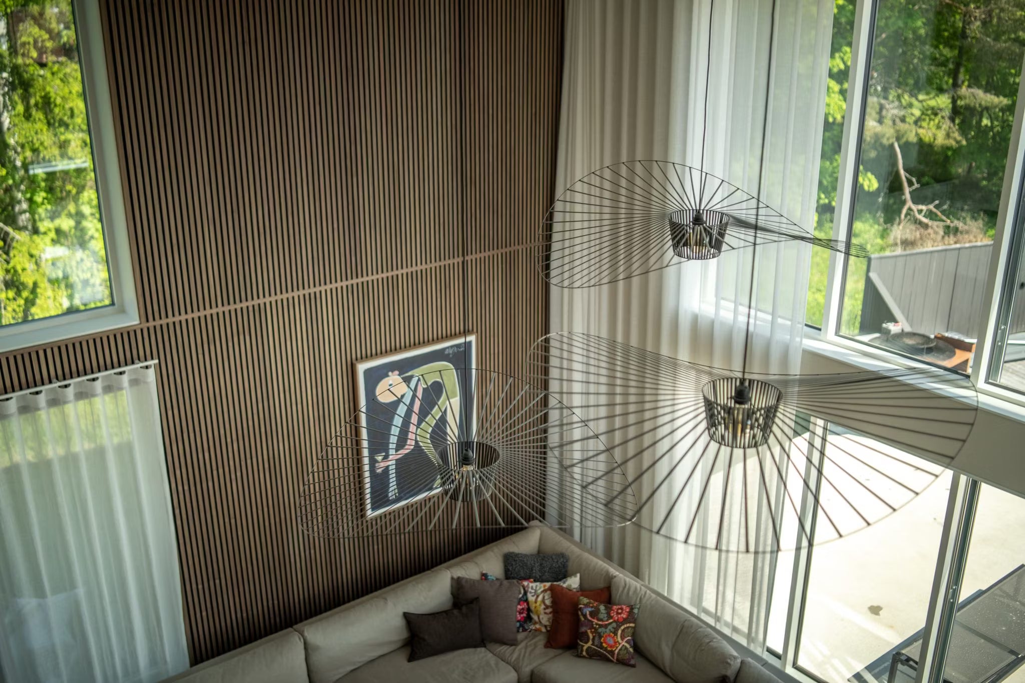 Moderno salón con paredes de Akupanel de madera nórdica, grandes ventanales, paneles acústicos abstractos, un sofá seccional y lámparas minimalistas de estructura metálica, visto desde arriba.