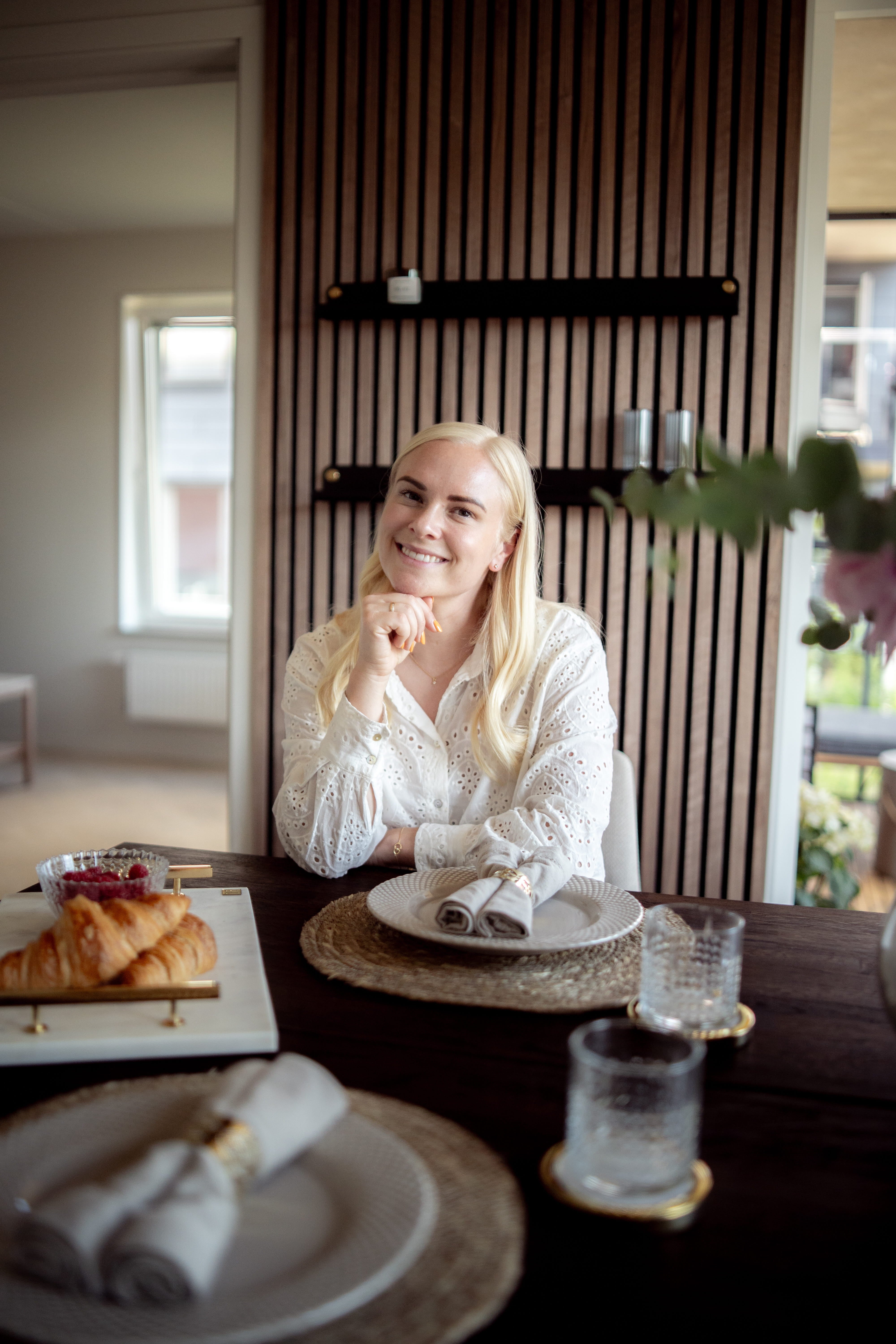 Una mujer de larga melena rubia se sienta en una mesa de comedor preparada para desayunar con cruasanes y cristalería, junto a un panel acústico nórdico en la pared.