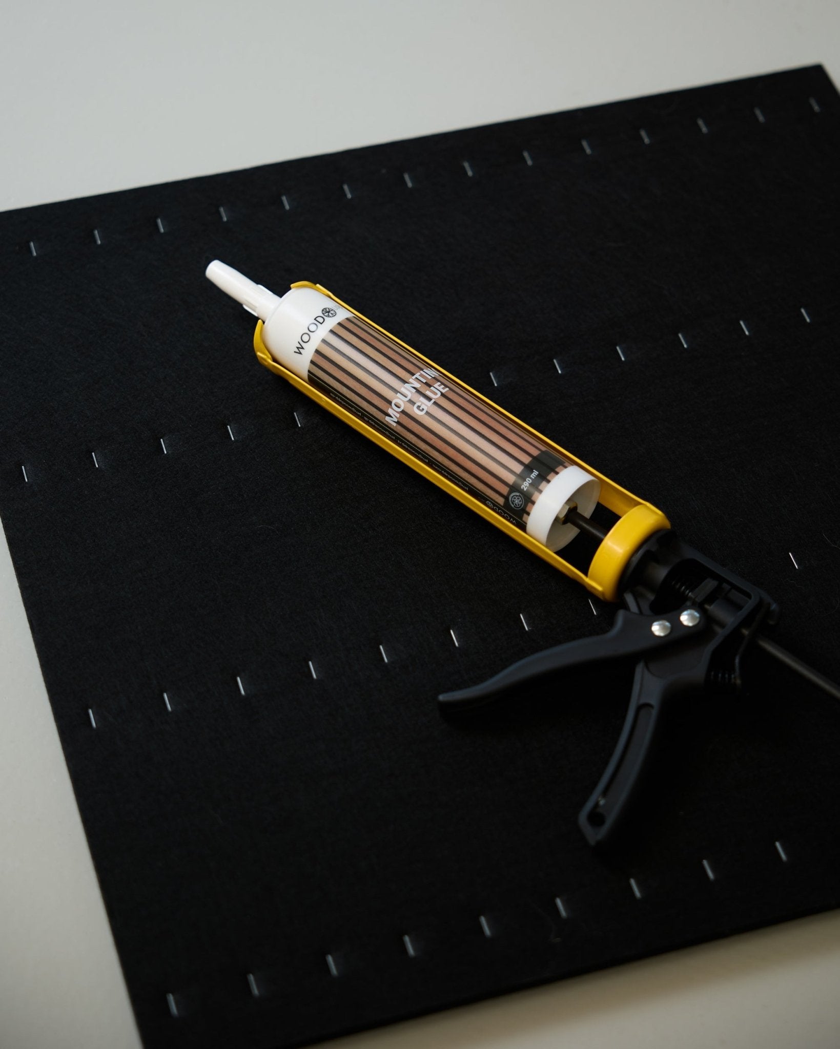 Una WoodUpp Grout Gun (290 ml) amarilla y black descansa sobre una alfombrilla black con una cuadrícula de grapas blancas, y se utiliza para instalar paneles acústicos.
