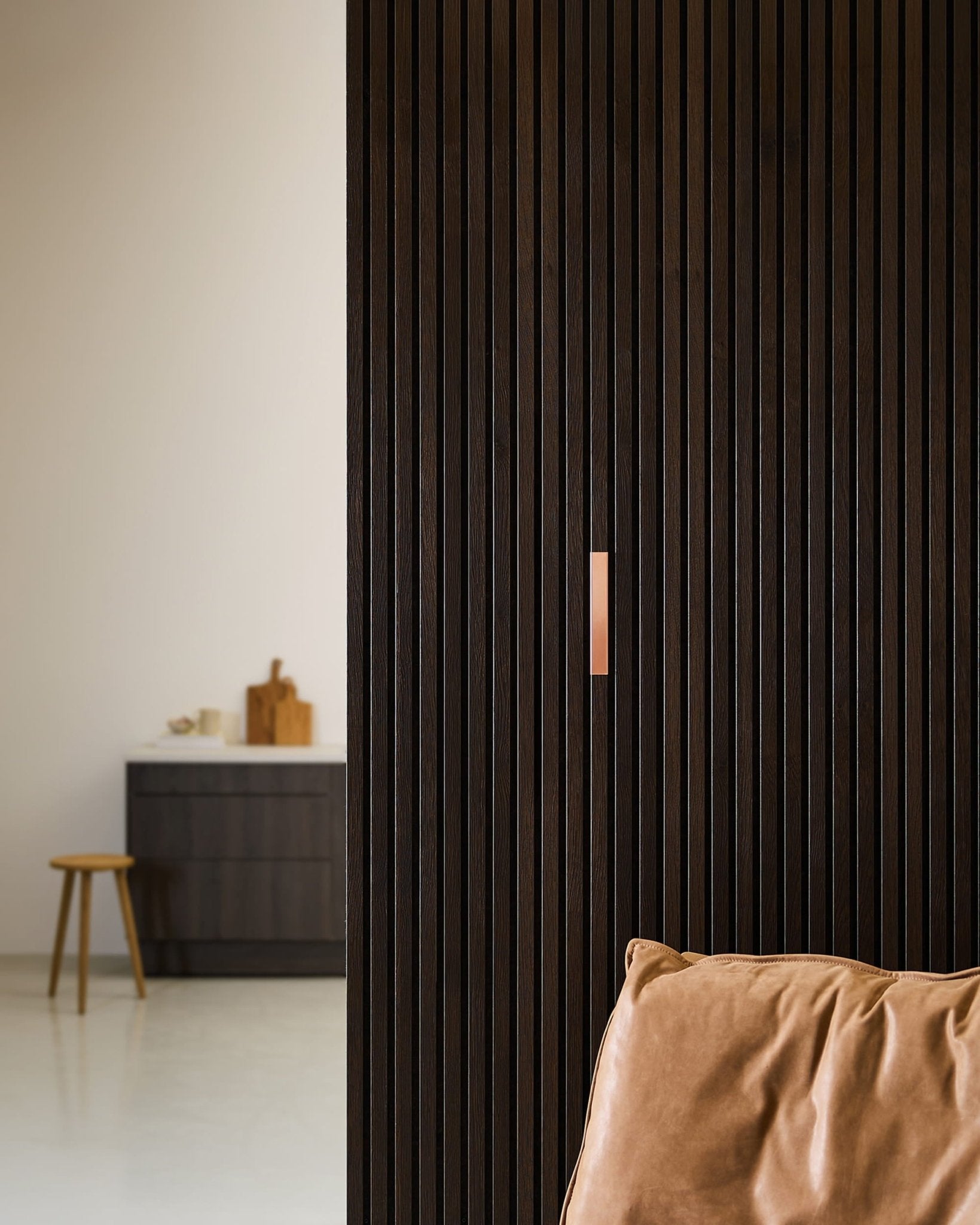 Interior minimalista con un sillón de cuero marrón, WoodUpp Liva | Cover / Paneles acústicos de cobre de 20 cm en la pared, y un taburete junto a una barra con tablas de cortar.