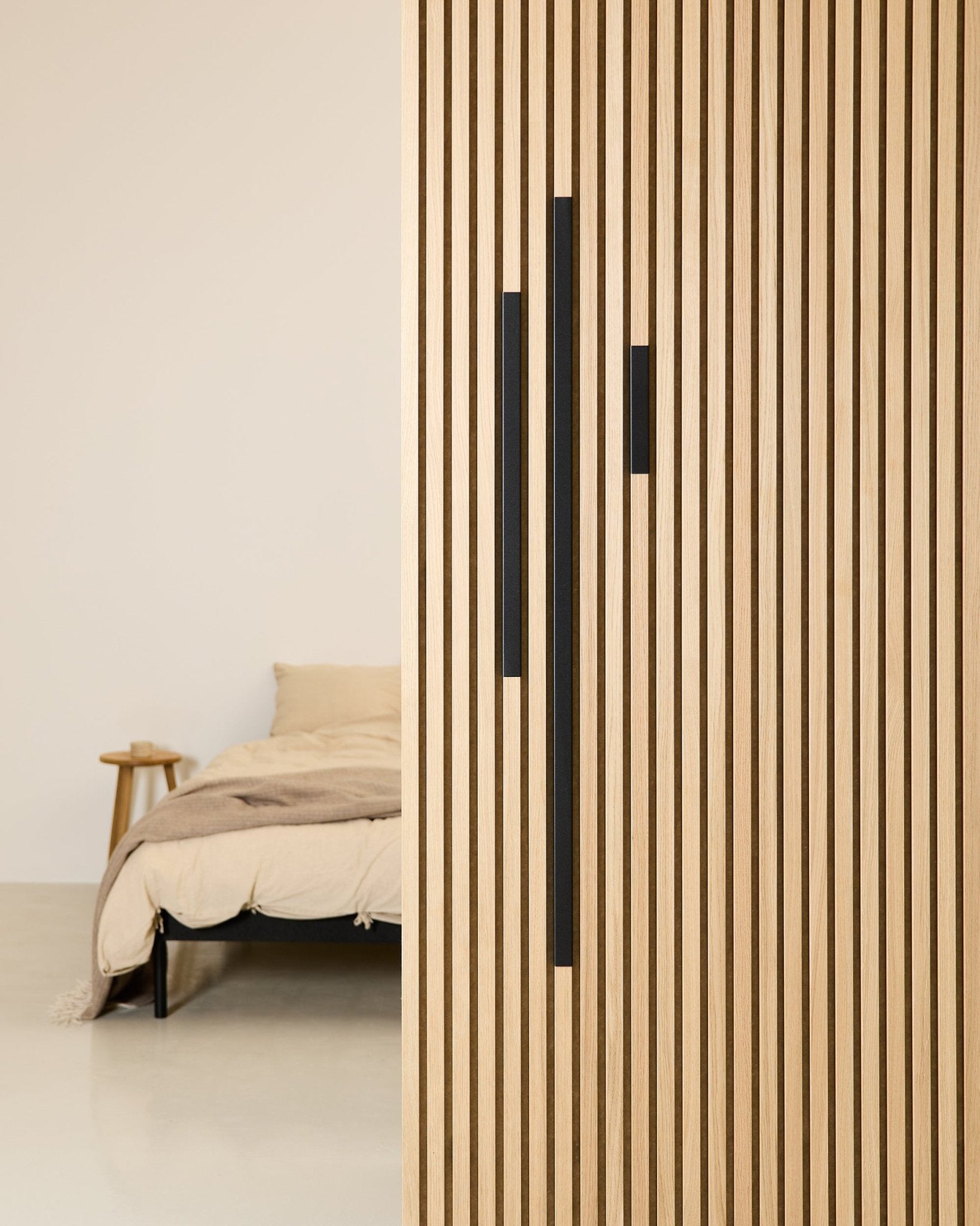 Dormitorio minimalista con una cama y una manta beige, parcialmente ocultas por un separador WoodUpp Akupanel de 60 cm en aluminio Black, de la serie Liva Cover.