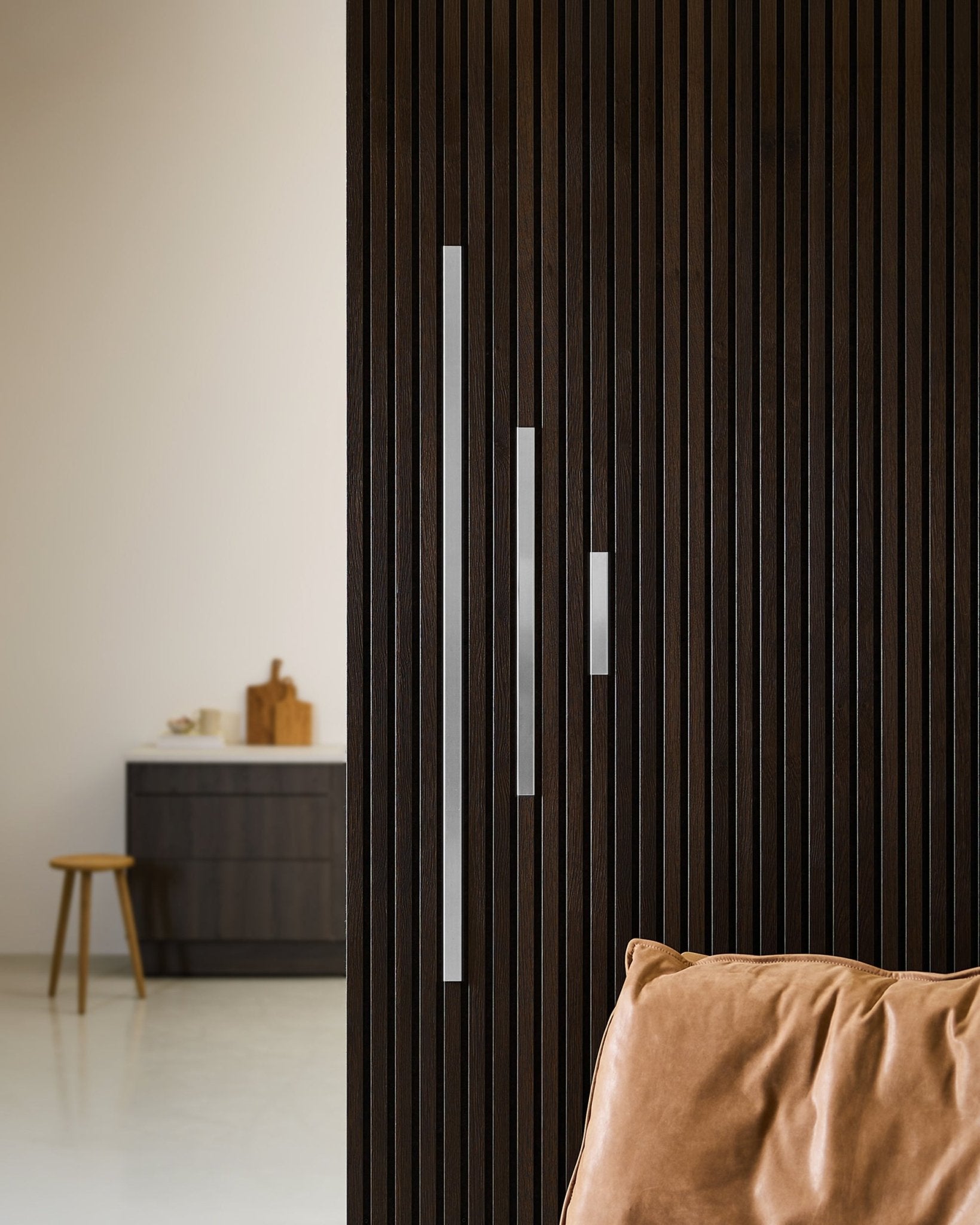 Interior minimalista con una pared Panel acústico de Akupanel en madera oscura, cover de WoodUpp Liva en steel pulido (20 cm), cojín color canela y mobiliario moderno.