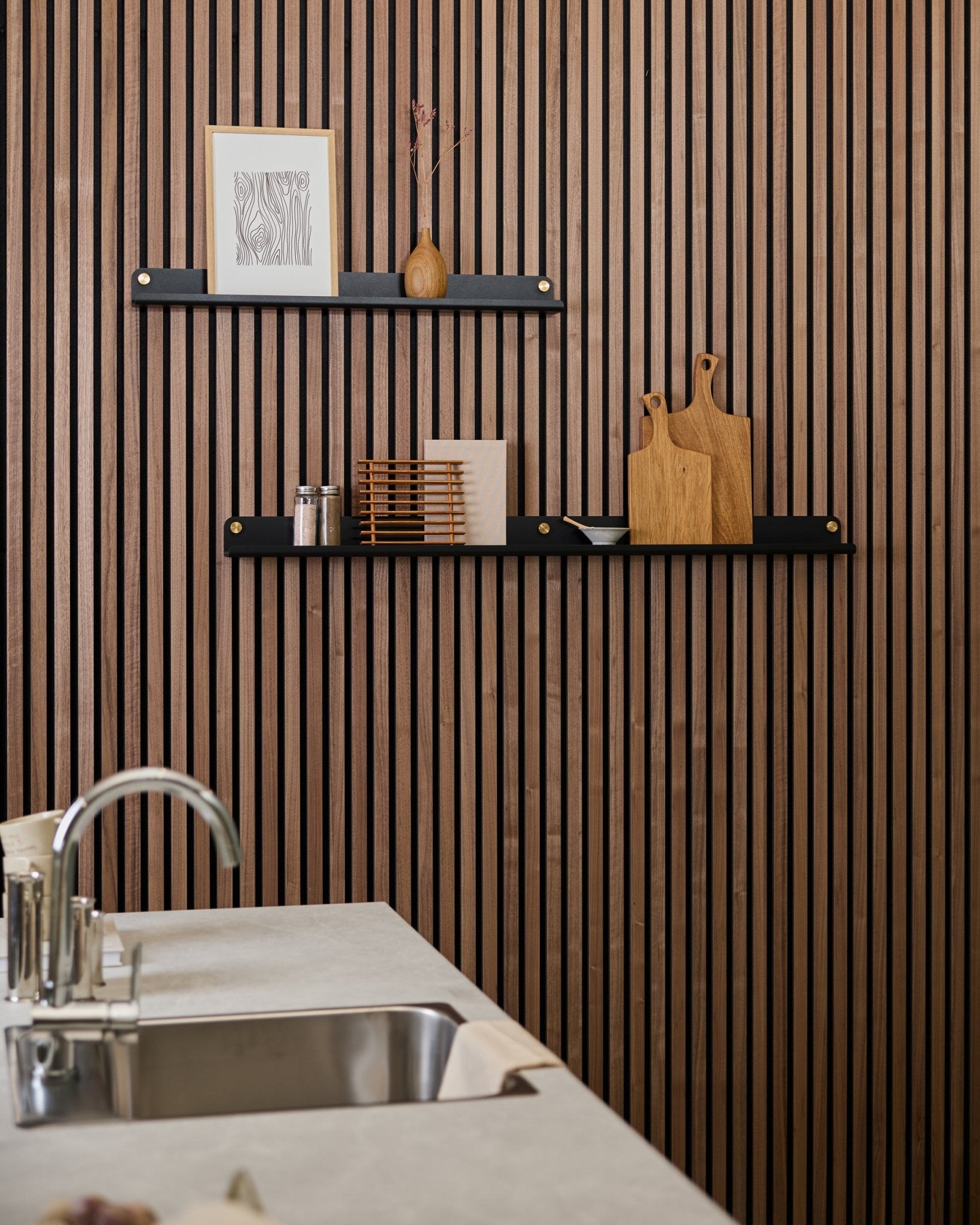 Cocina minimalista con una pared acústica de lamas de madera en vertical Akupanel, estantes black, tablas de cortar, objetos decorativos small y una encimera con fregadero moderna delante.