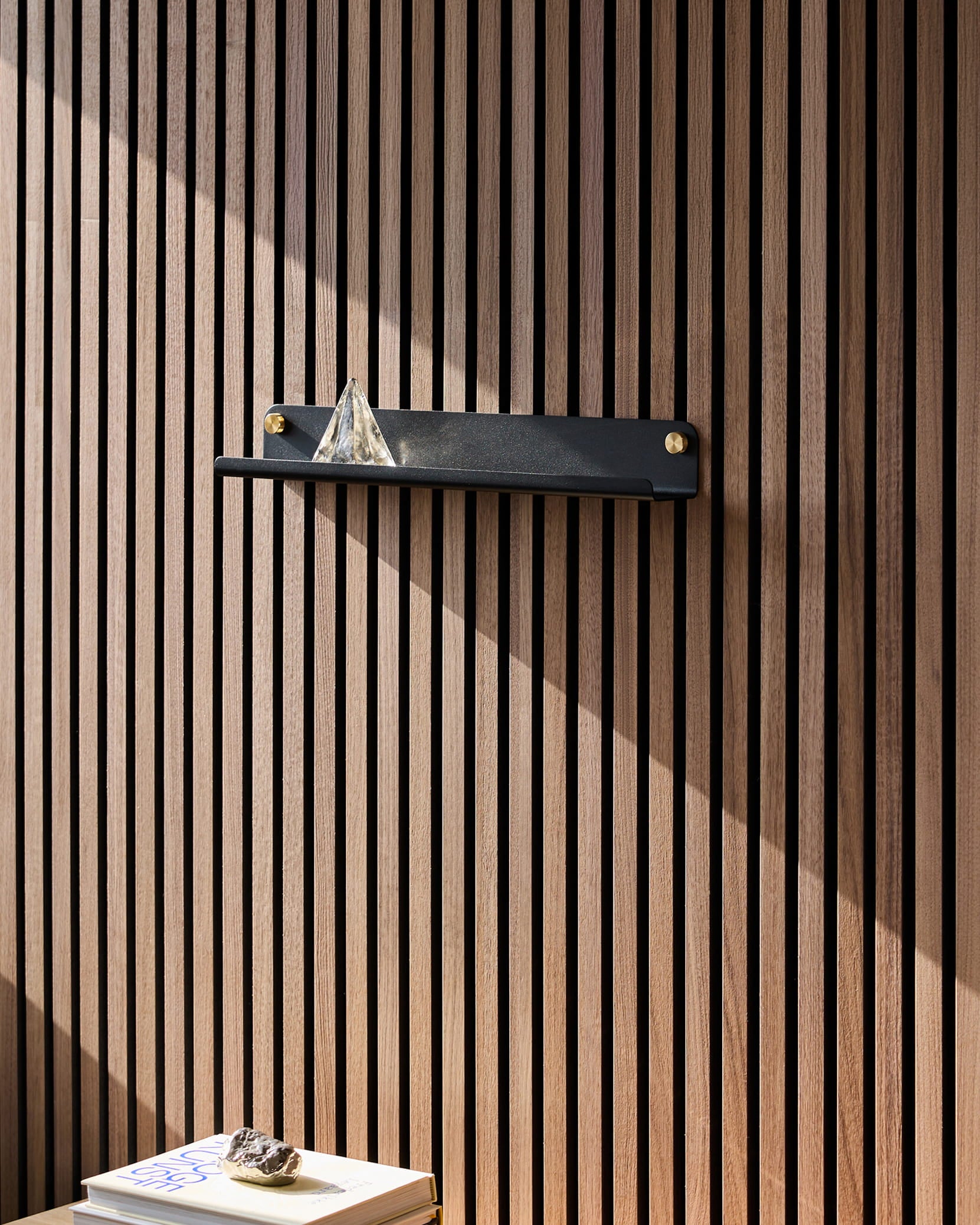 Una estantería estrecha Norr de color negro, de la marca WoodUpp, colocada sobre un panel acústico de madera Akupanel, sostiene una pirámide de cristal; la luz del sol incide sobre los libros que hay debajo, coronados por una piedra.
