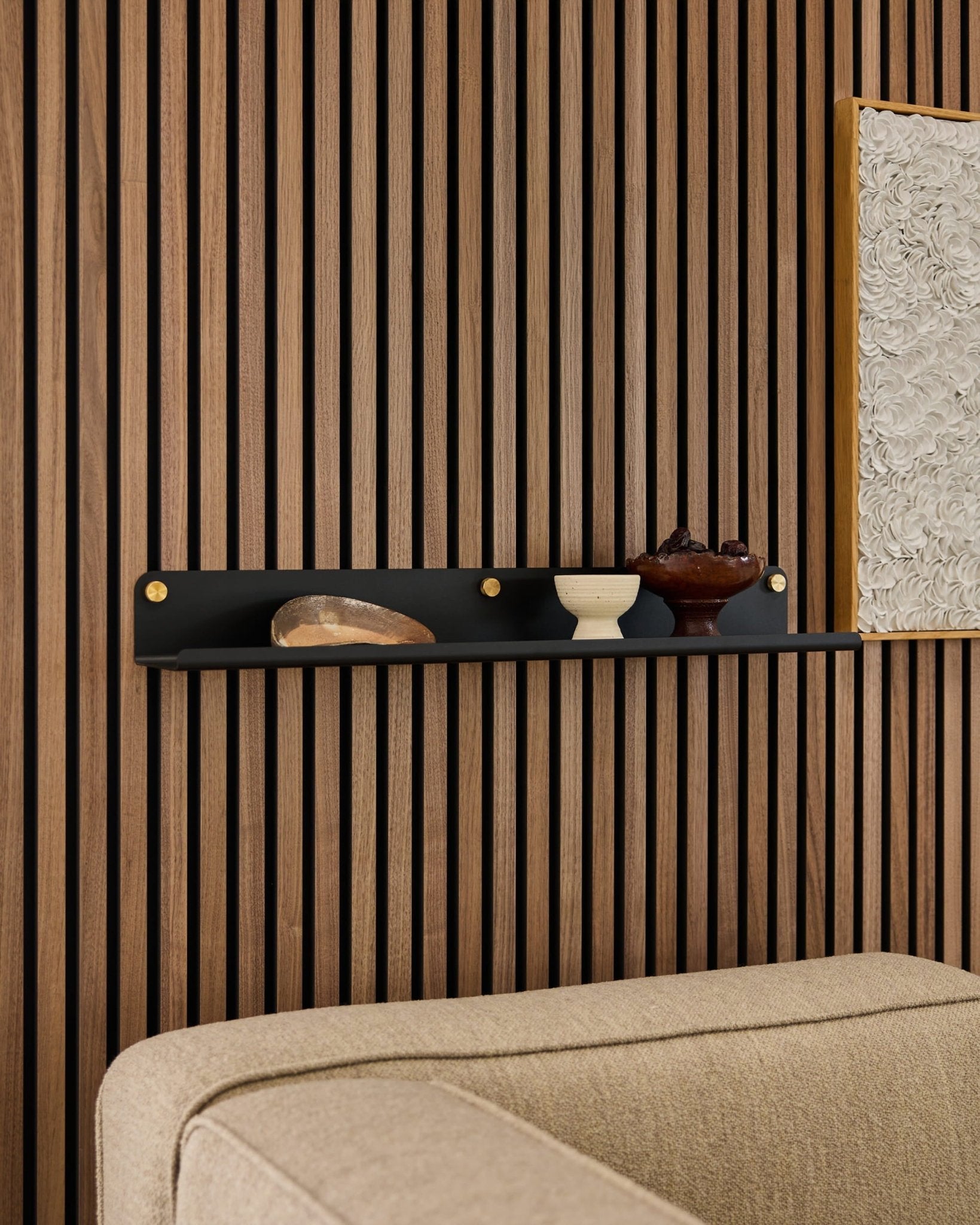 Estantería ancha WoodUpp Norr en negro (75,9 cm) con elementos decorativos sobre un sofá beige, montada sobre un moderno panel de pared acústico de listones de madera.