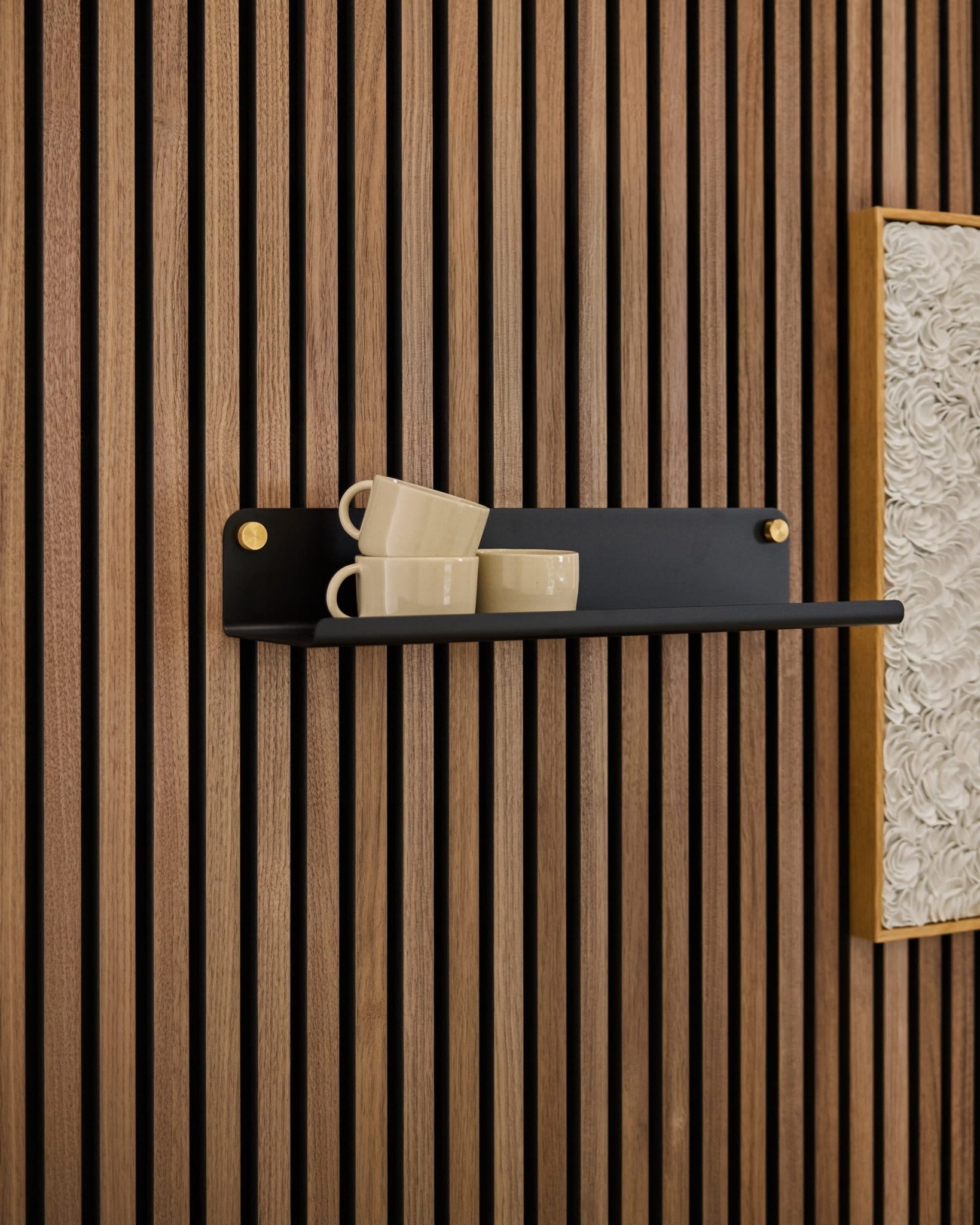 Una estantería Norr Wide de color negro, de la marca WoodUpp, sostiene tres tazas de color beige, colocadas sobre un panel acústico Akupanel junto a una obra de arte enmarcada de color blanco con textura.