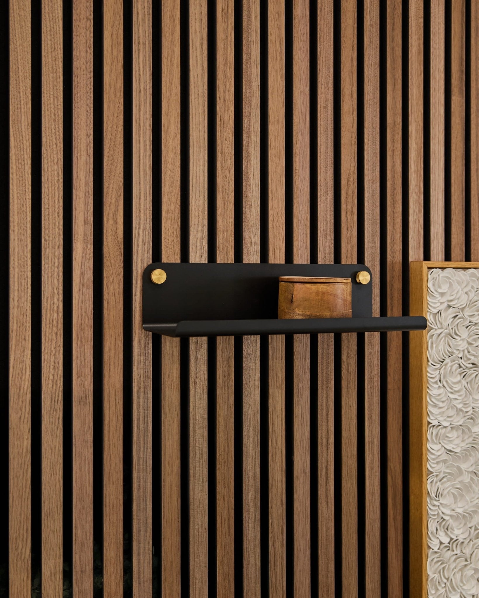 Una pequeña caja de madera colocada sobre una estantería WoodUpp Norr Wide (negra, 35,9 cm) fijada a unos listones acústicos verticales de madera Akupanel, con un panel blanco texturizado a la vista.