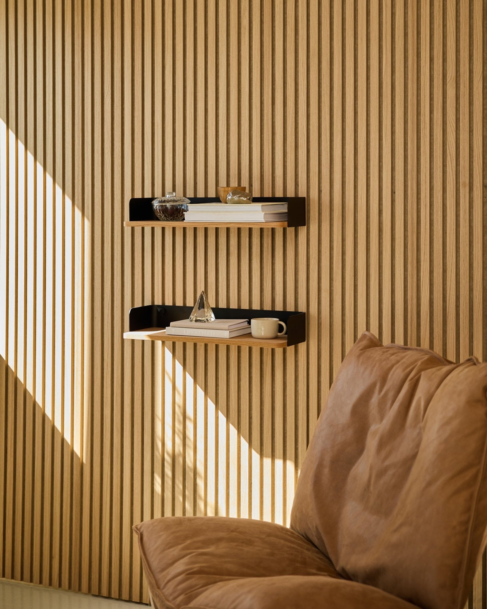 Silla Brown junto a la estantería Linn Bookshelf de WoodUpp en Natural Oak, con libros y objetos decorativos dispuestos con luz natural junto a un panel de listones de madera nórdico para pared Panel acústico.