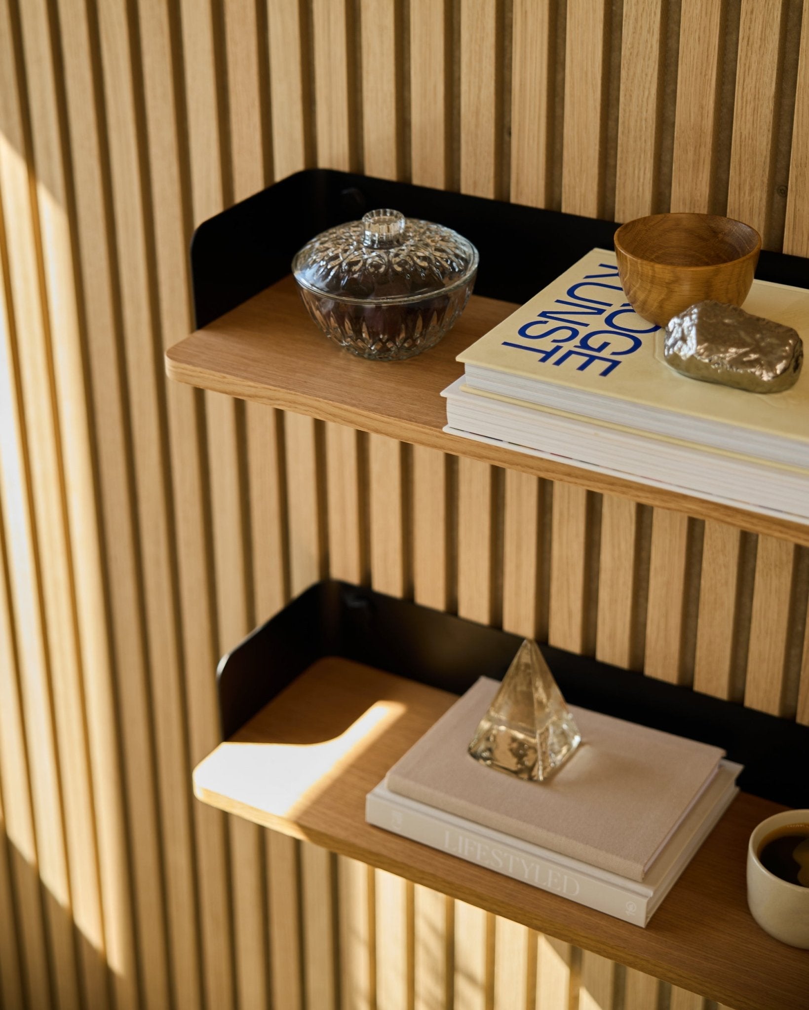 La estantería Linn Bookshelf de WoodUpp en Natural Oak (55,9 cm) sostiene un tarro de cristal, una taza, una pirámide dorada y libros sobre paneles acústicos de madera Akupanel a la luz del sol.
