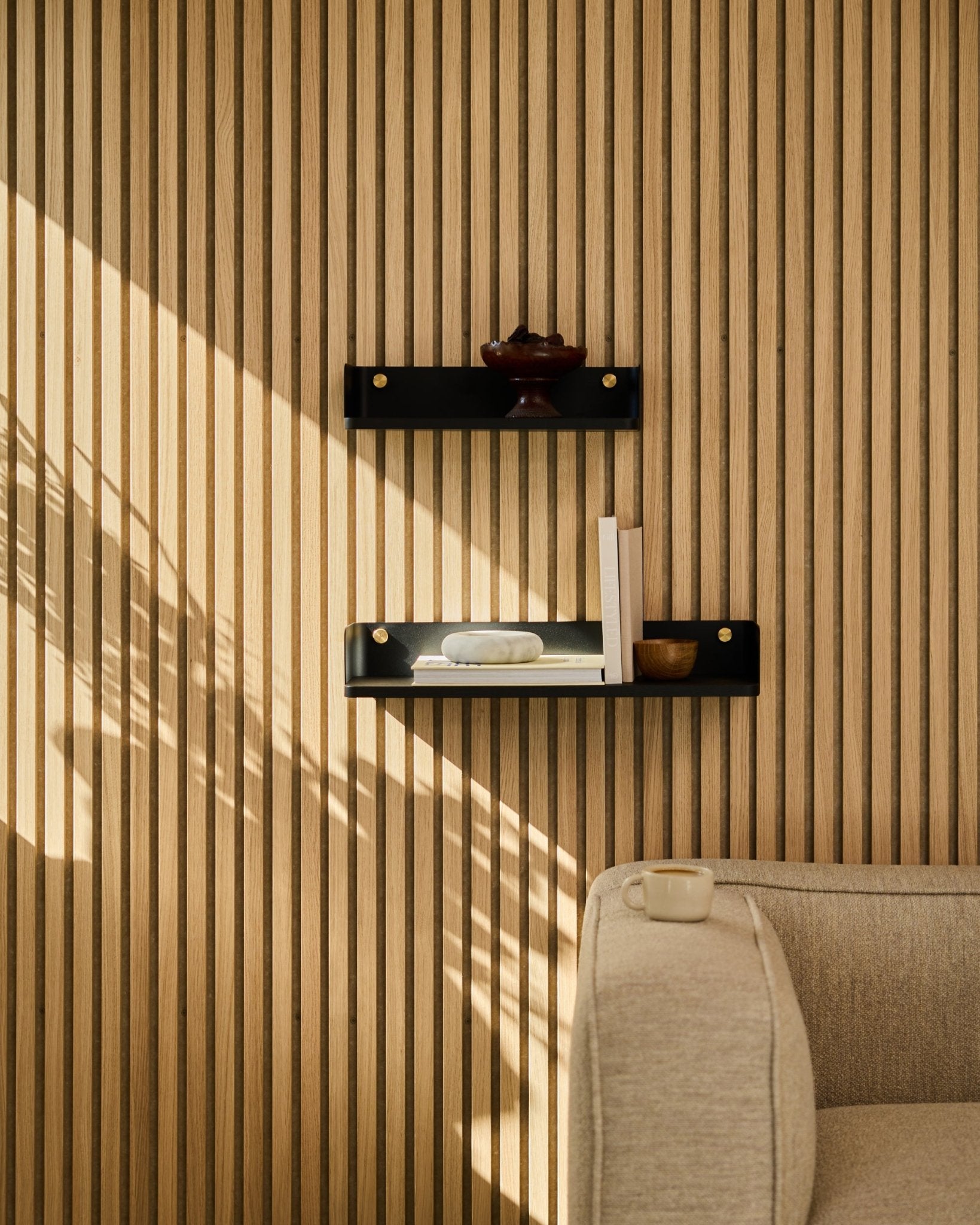 La luz del sol ilumina un sofá beige y la estantería Linn de WoodUpps en black oak, 55,9 cm, colocados frente a un panel de listones nórdico minimalista Panel acústico.