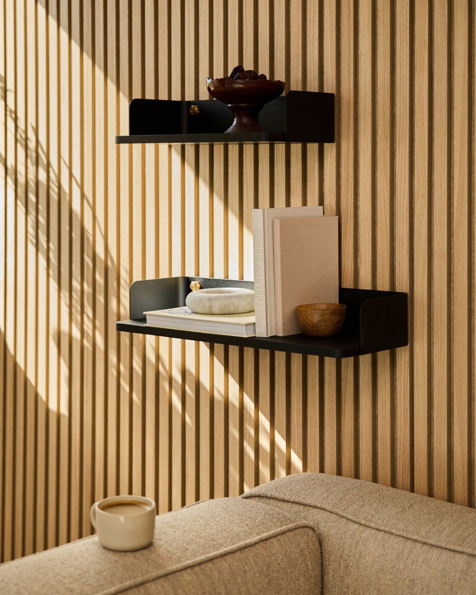Una estantería WoodUpp black oak (55,9 cm) muestra libros, objetos decorativos y un cuenco sobre una pared con panel acústico de madera; en primer plano, un sofá beige y una taza.