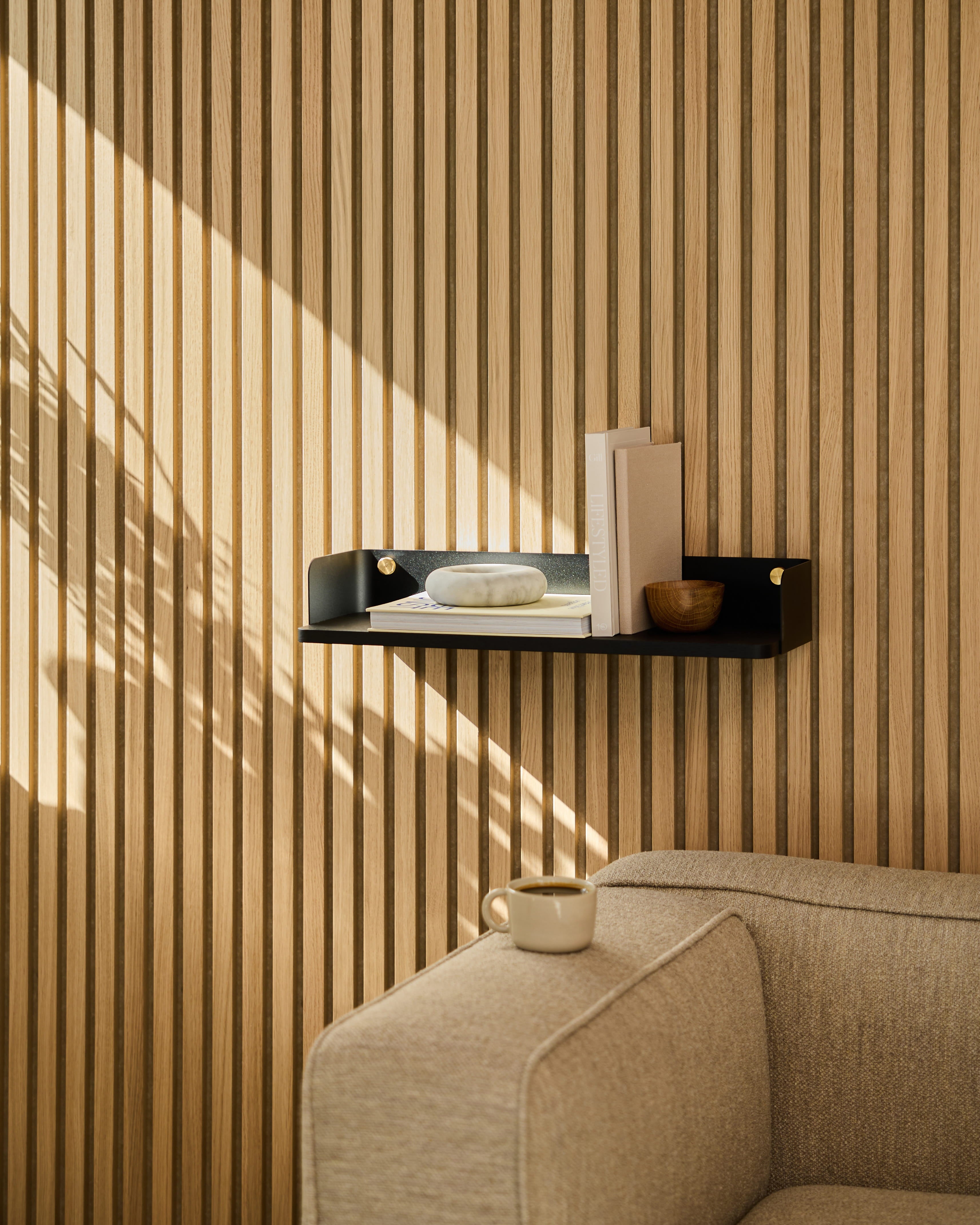 Una estantería A Linn | Bookshelf / Black Oak de WoodUpp decora la pared situada encima de un sofá beige. La luz del sol proyecta sombras sobre la pared revestida con paneles acústicos de madera a rayas.