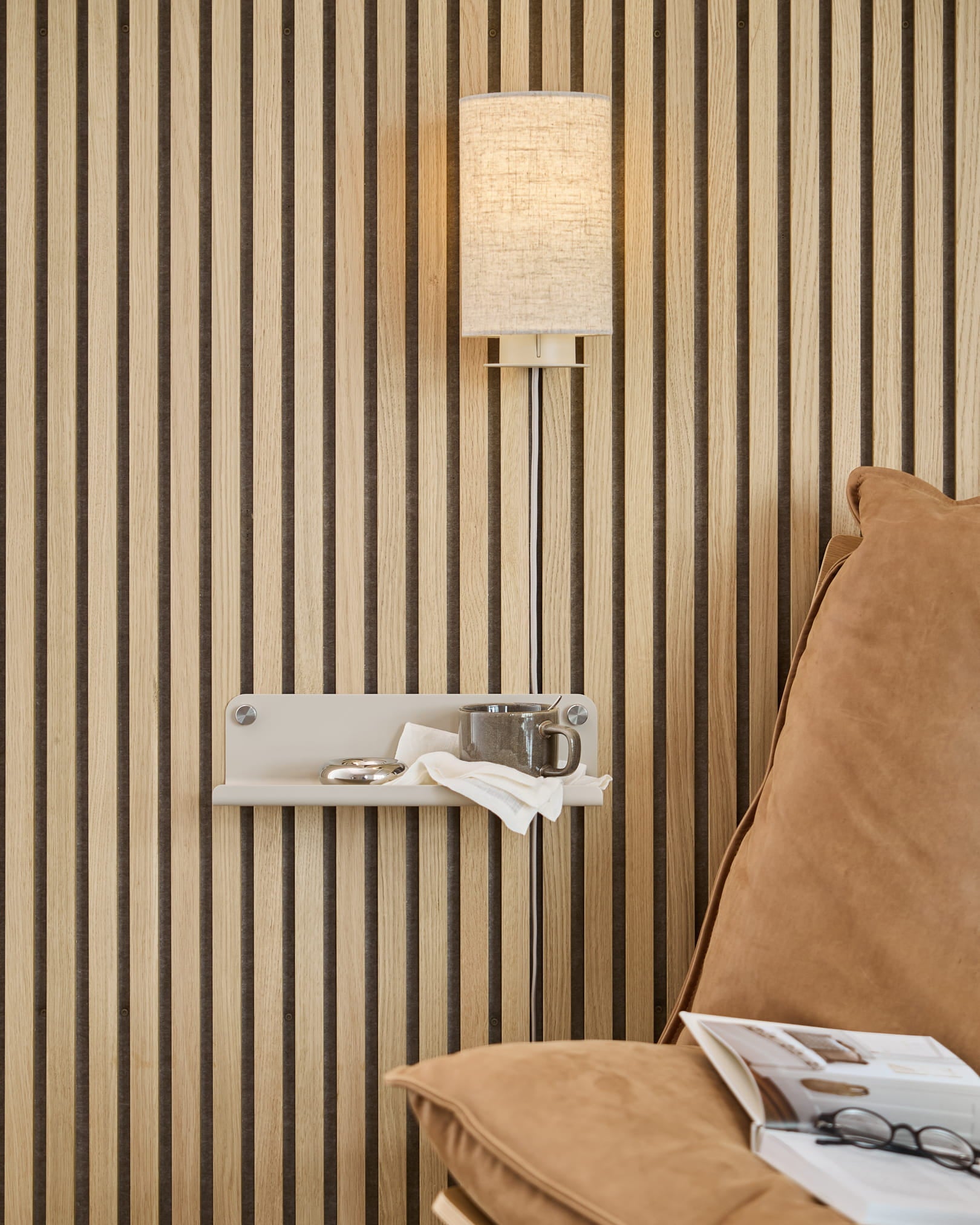 Pared acústica vertical de listones de madera «Akupanel», aplique de pared beige, estante pequeño con una taza y una servilleta, sofá de color tostado con una revista abierta.