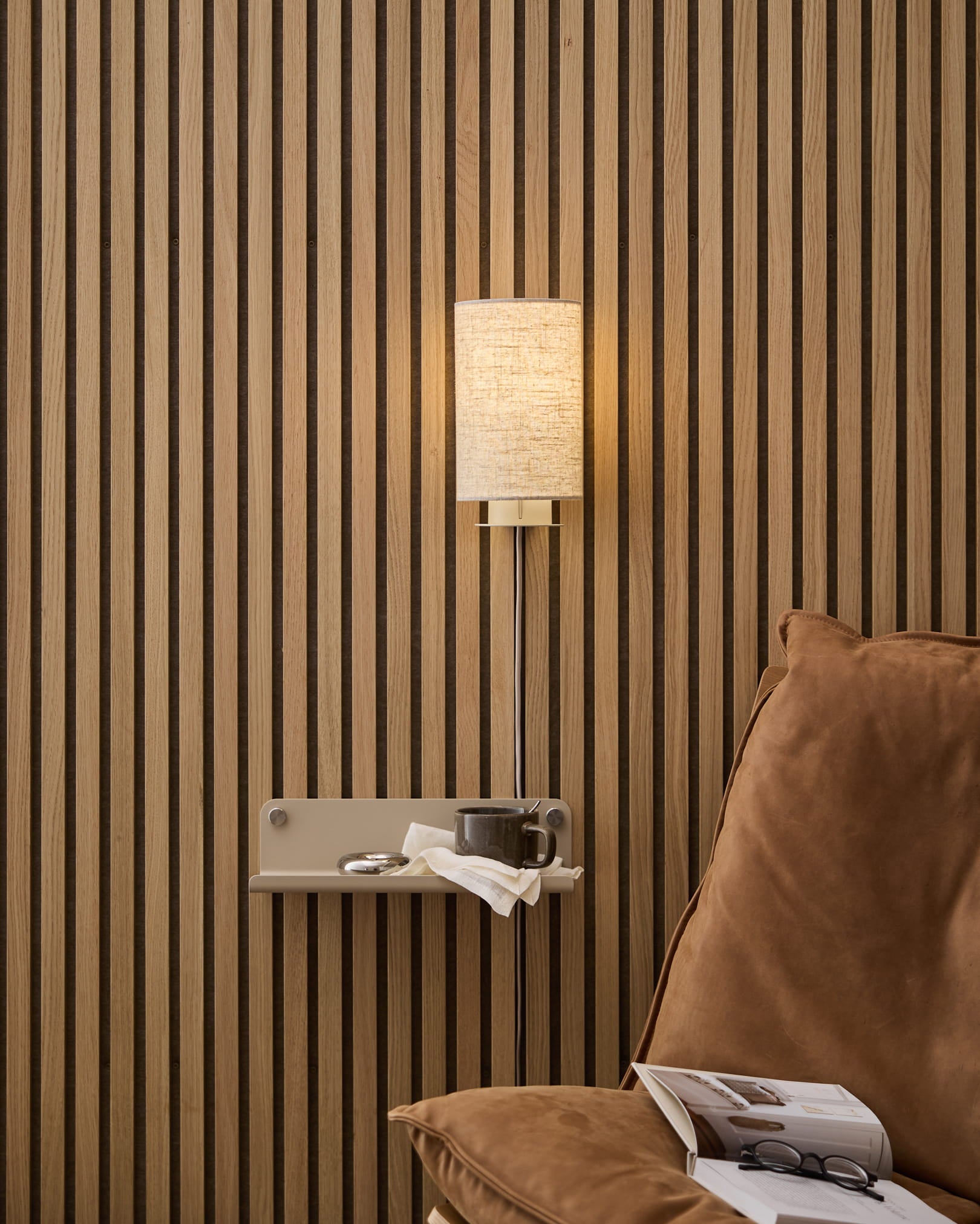 Una lámpara de pared Feya de WoodUpp en lino y algodón beige, montada sobre un acogedor sillón y una mesa auxiliar con libro y taza, frente a un panel nórdico de lamas de madera Panel acústico.