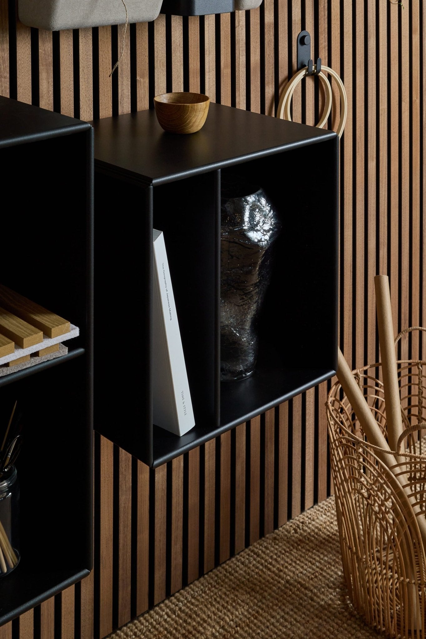 La estantería Arka / Negra de WoodUpp, una estantería modular con dos compartimentos, situada junto a una cesta de mimbre y un cuenco de madera sobre un panel acústico nórdico de listones.