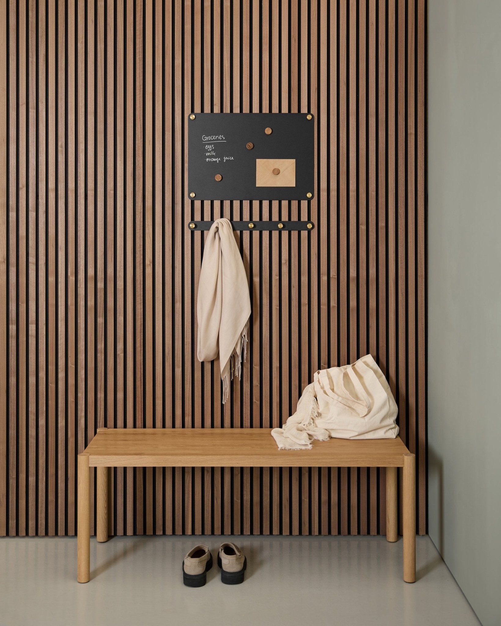 Recibidor minimalista con un banco de madera, dos bufandas, sandalias y una pizarra magnética WoodUpp Norr small sobre un panel acústico de listones de madera Akupanel.