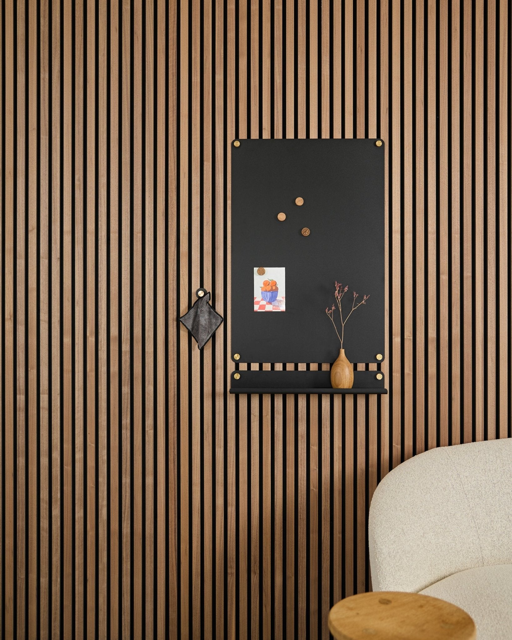 Un large black Norr tablero magnético de WoodUpp, diseñado para paneles acústicos, cuelga junto a una silla tapizada blanca con notas, un jarrón y un adorno.