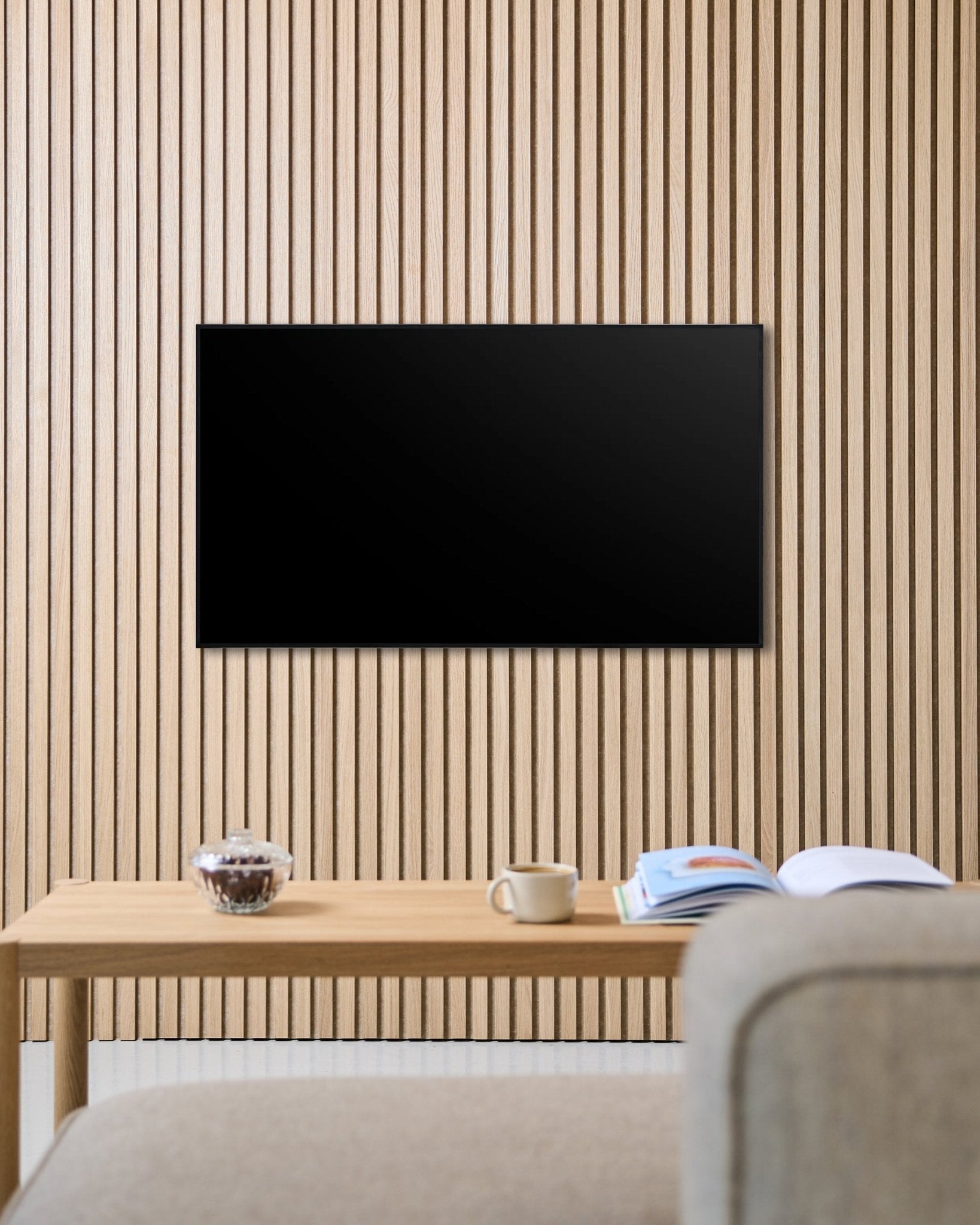 Un soporte de TV Fjell (small) de WoodUpp fija una pantalla plana a una pared acústica nórdica de lamas de madera Akupanel, sobre una mesa con una taza, un cuenco y una revista abierta junto a un sofá beige.