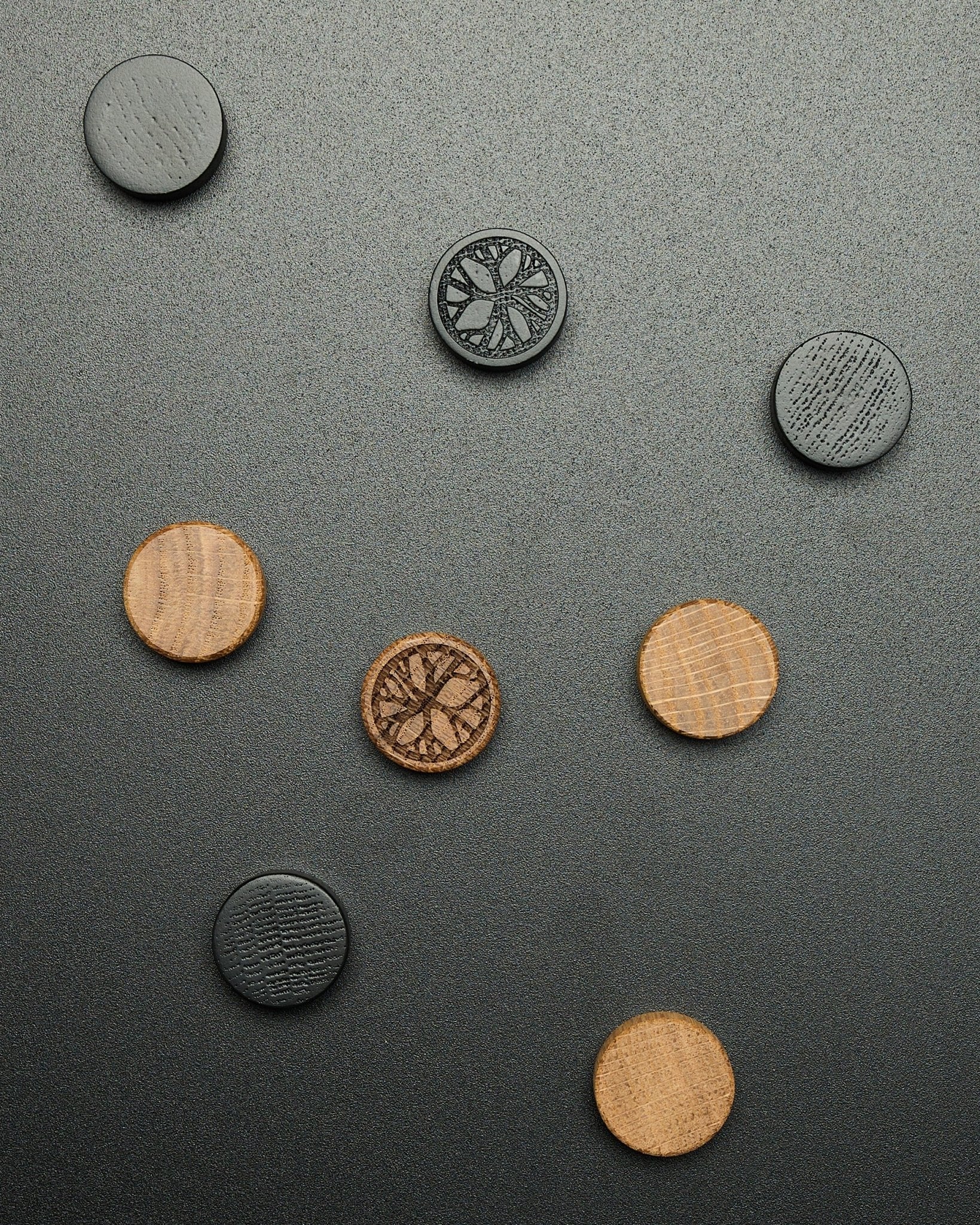Cuatro imanes redondos black oak del juego de imanes WoodUpp Norr, dispuestos de forma suelta sobre una superficie oscura con textura, diseñados como accesorios Akupanel.
