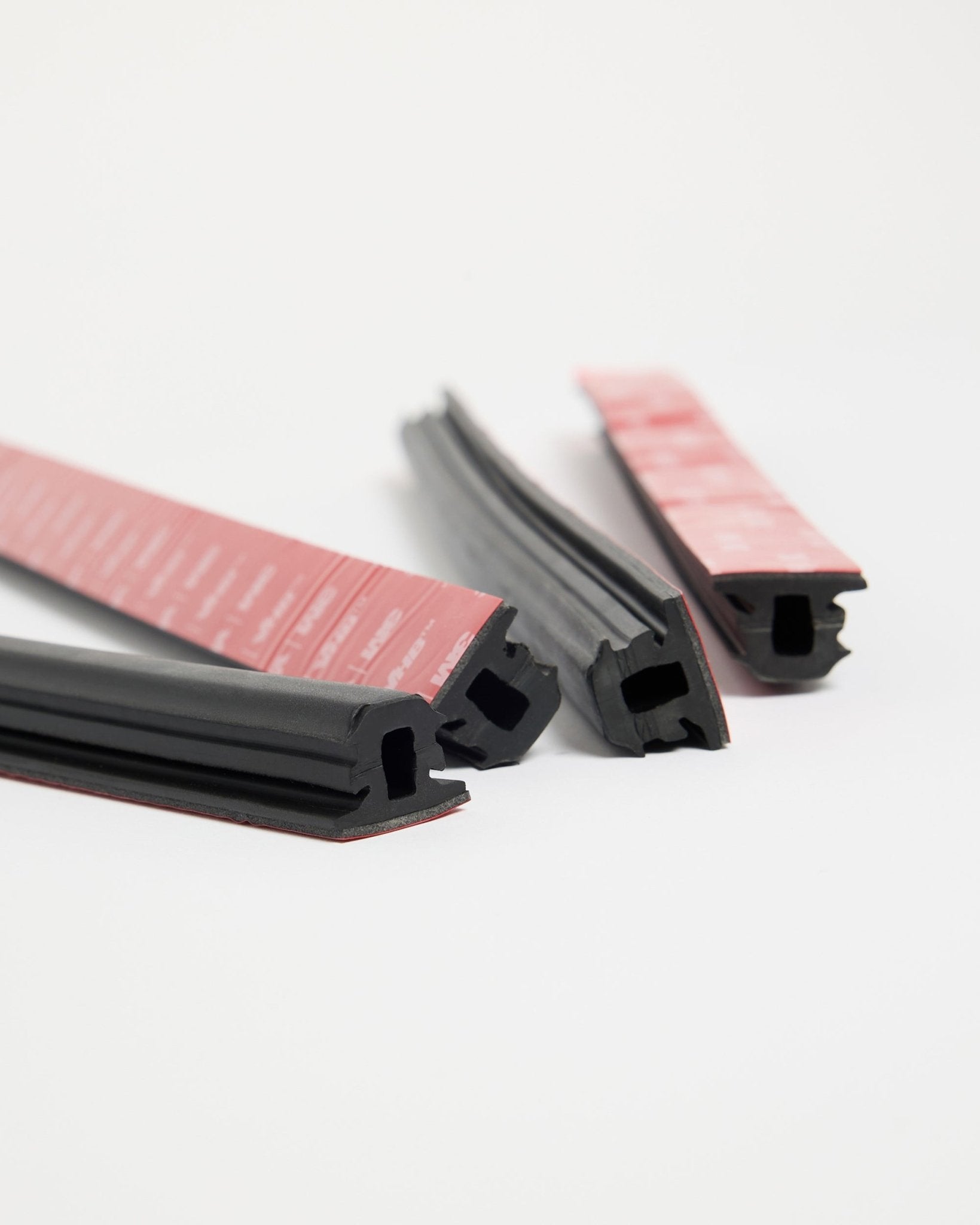 Cuatro tiras de goma Pop-on Profile Dupps black de WoodUpp, con adhesivo rojo, diseñadas para una instalación Akupanel, dispuestas en varias direcciones sobre una superficie blanca.