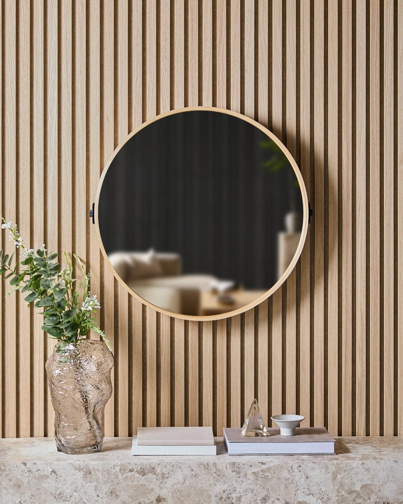 El Linn | Mirror en Natural Oak de WoodUpp está montado sobre un Panel acústico nórdico de listones, encima de una balda de piedra con un jarrón de vidrio, libros y objetos decorativos.