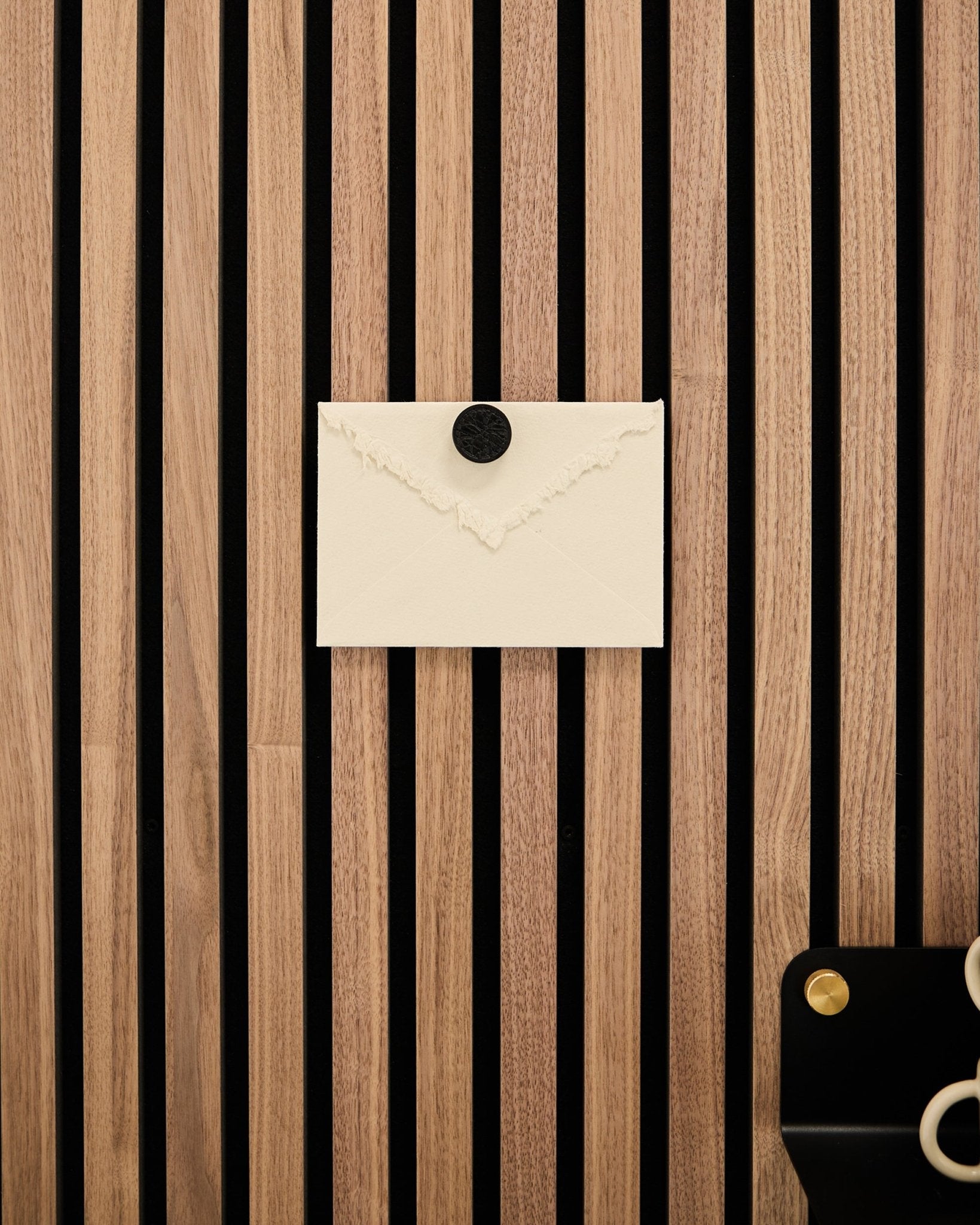 Un sobre color crema está sujeto a un panel acústico de lamas de roble Akupanel de WoodUpp Panel acústico con el juego de imanes para póster Norr en Black Oak; unas tijeras descansan en la parte inferior derecha.