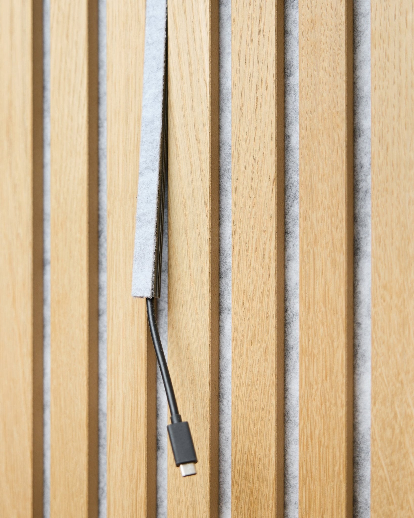 Un cable USB-C black pasa por un grey Pop-on Cable Cover de WoodUpp Dupps entre listones acústicos de madera vertical con respaldo de fieltro grey.