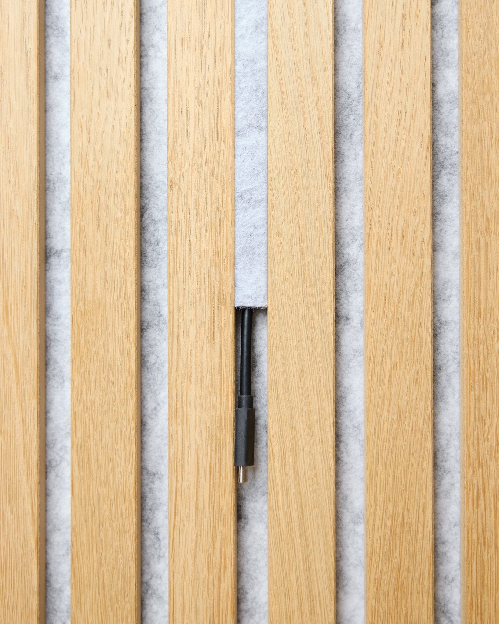 El juego de Pop-on Cable Cover Dupps (grey, 3 uds.) de WoodUpp organiza los cables entre listones acústicos de madera clara Akupanel sobre un fondo blanco con textura.