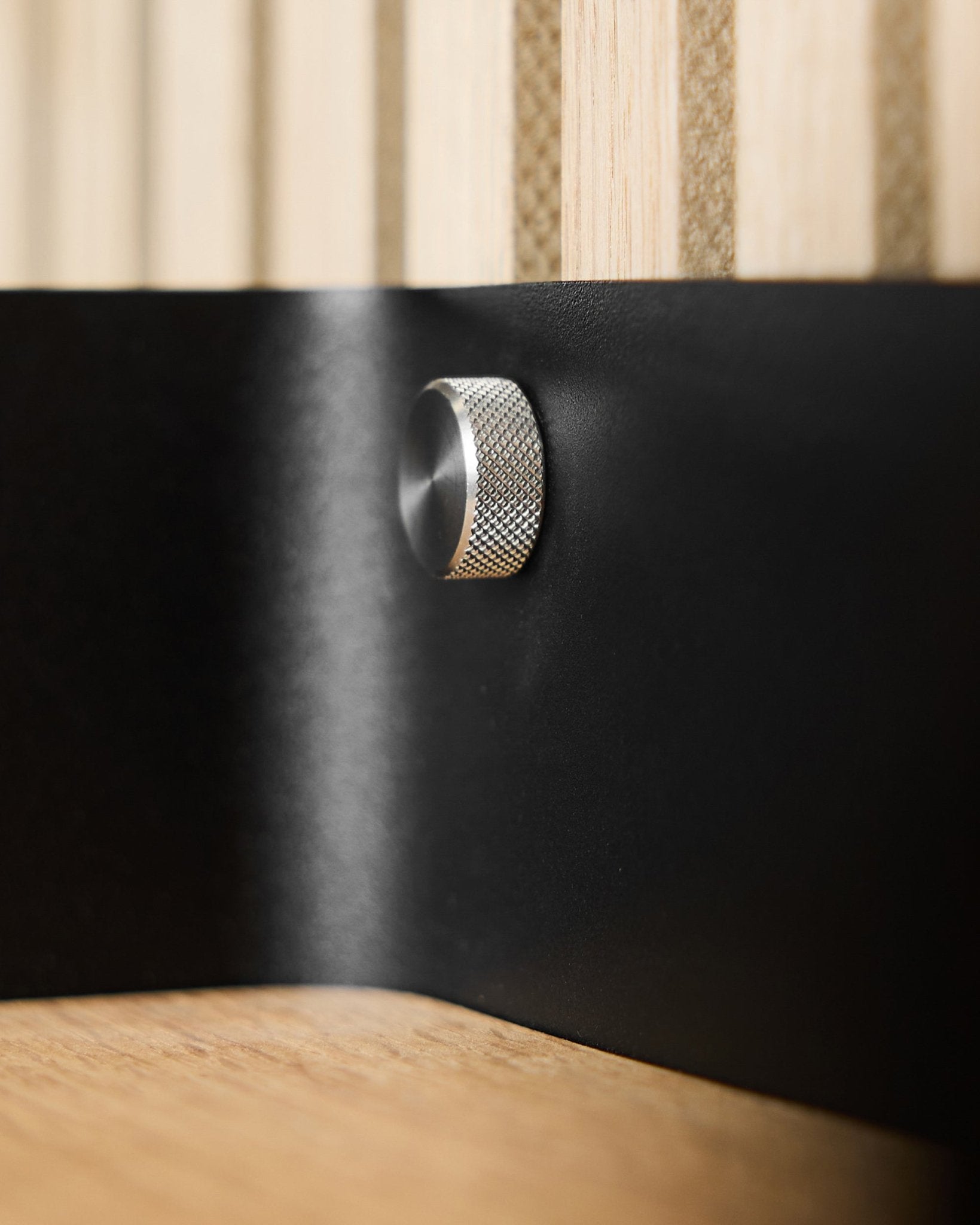 Primer plano de dos tornillos de apriete manual WoodUpp (M5x10 mm, acero inoxidable steel) sobre una superficie metálica black, con lamas acústicas de madera Akupanel al fondo.