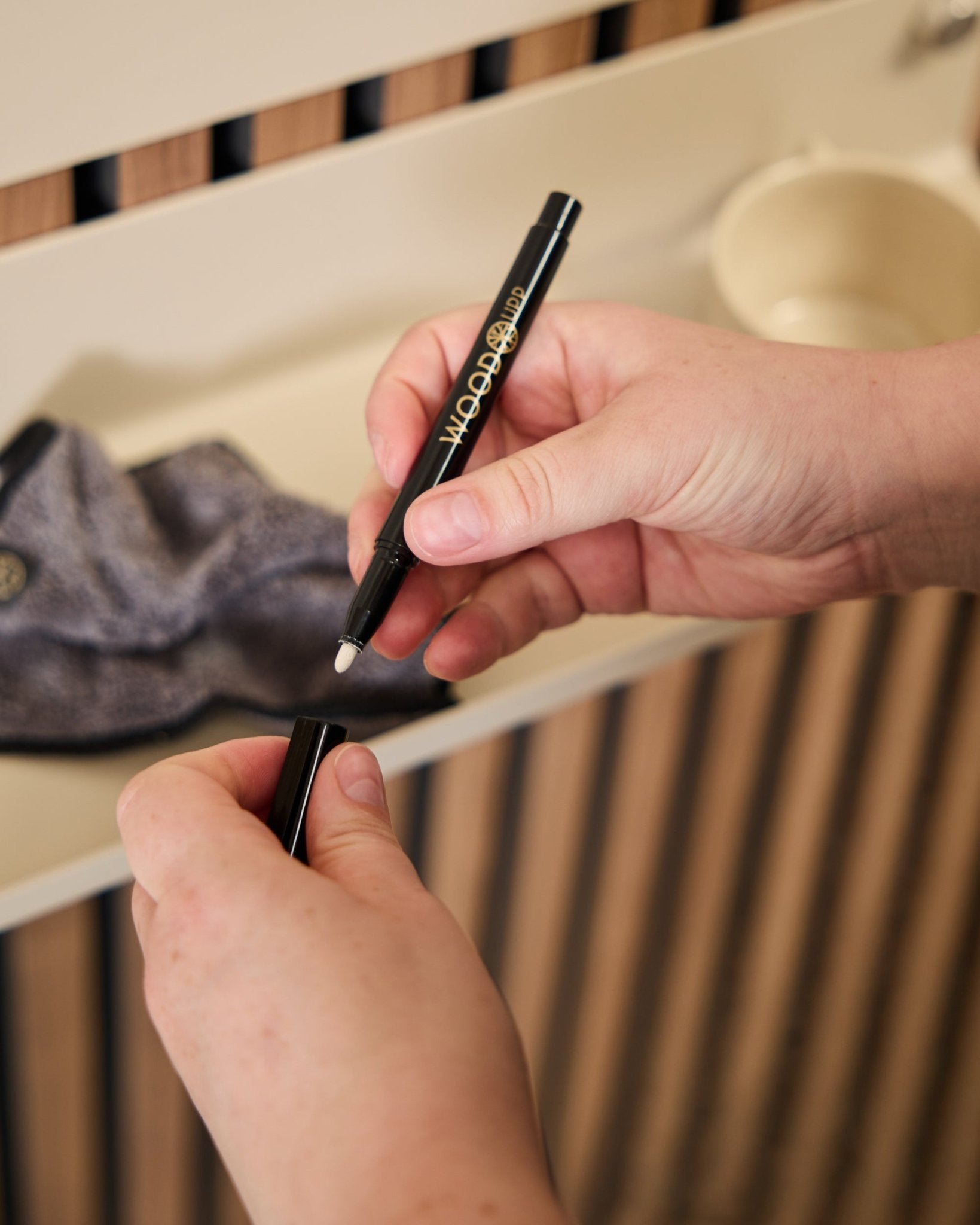 Una persona sostiene un rotulador WoodUpp Norr | Blackboard set abierto junto a un paño de microfibra y una taza beige, productos diseñados para usarse con paneles acústicos de madera.
