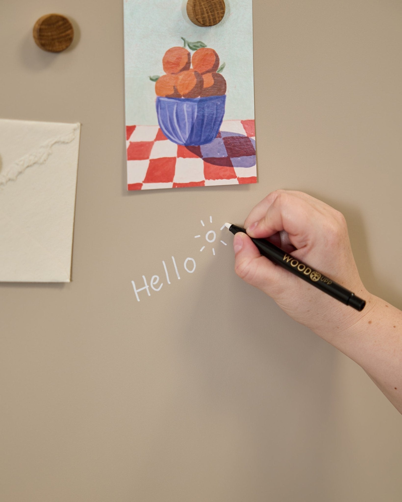 Una mano escribe hello con un dibujo de un sol en la Blackboard Norr de WoodUpp, colocada junto a unas ilustraciones sujetas con imanes redondos sobre un Panel acústico.