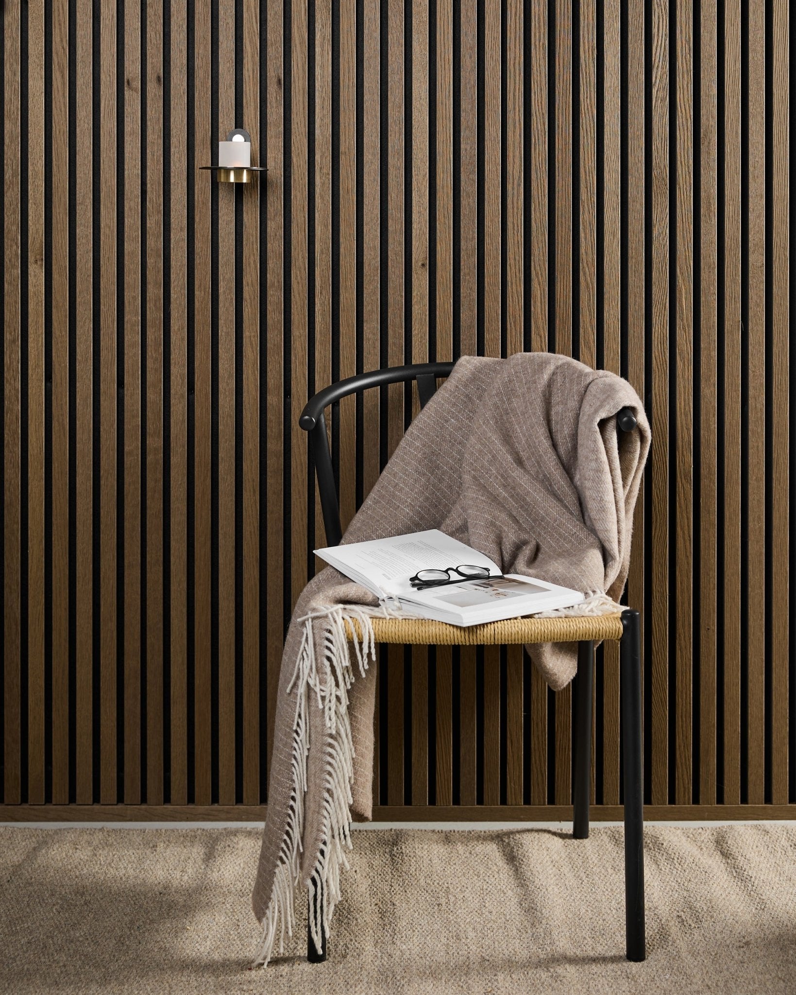 Una silla moderna con una manta beige, un libro abierto y unas gafas se sitúa junto a un panel acústico de madera Akupanel y un portavelas WoodUpp Norr en black y brass.
