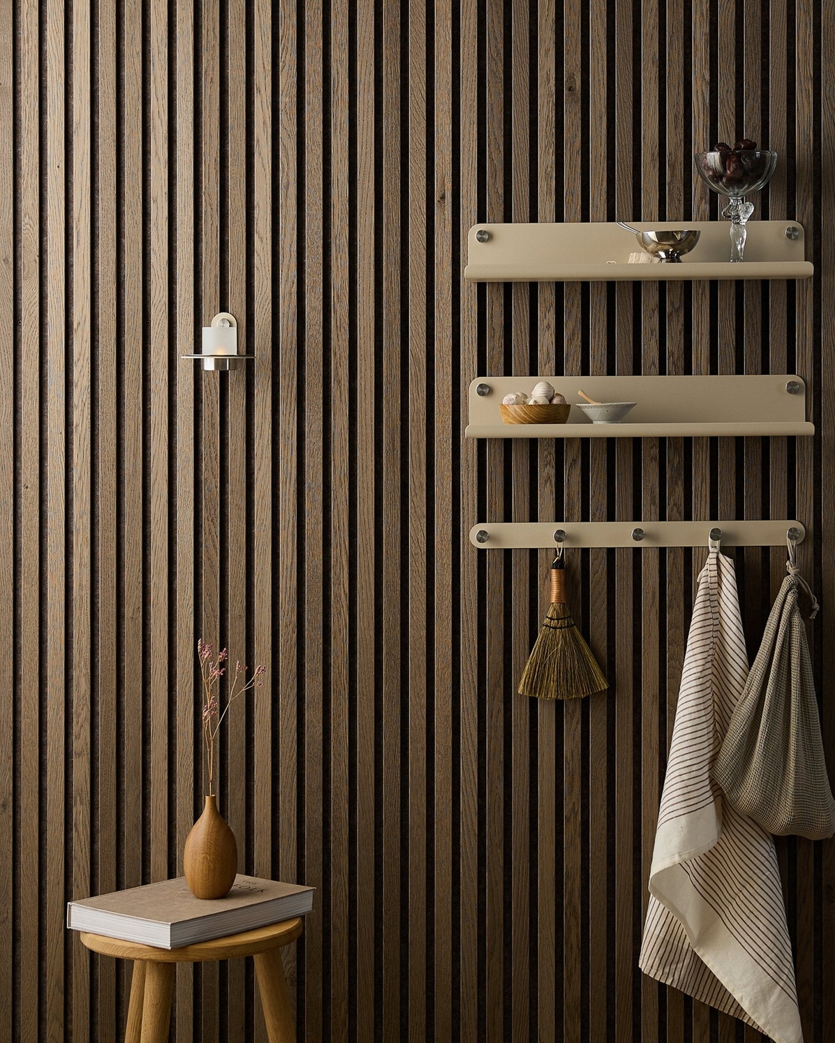 Un taburete de madera con flores secas está junto a un Panel acústico Akupanel de WoodUpp, al lado de unas estanterías beige con vajilla, un cepillo, toallas y un portavelas Norr.
