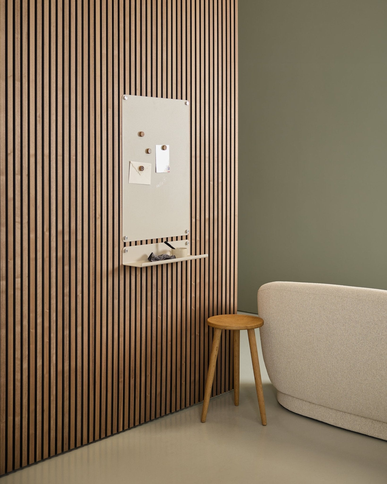 Interior minimalista con sofá beige y taburete de madera, con un tablero magnético WoodUpp Norr large beige montado en un panel acústico de madera Nordic Akupanel junto a una pared verde.