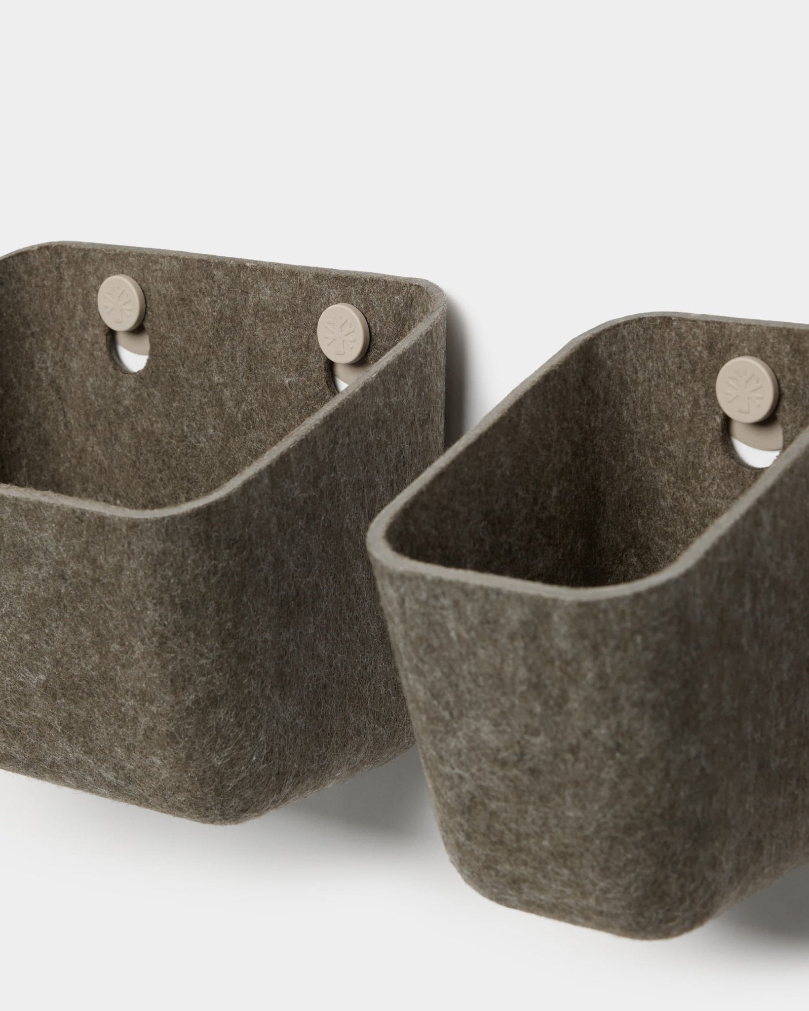 Dos recipientes de fieltro Luva, en gris oscuro cálido, de la marca WoodUpp, cuelgan de ganchos de color beige fijados a un panel acústico, lo que ofrece un sistema de almacenamiento de pared minimalista con esquinas redondeadas.