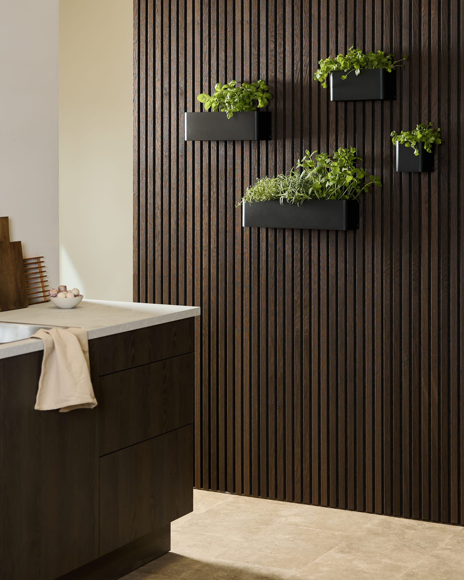 Cocina moderna con un Panel acústico de madera oscura WoodUpp Akupanel, una Plant Box Norr black con hierbas y muebles oscuros rematados con una encimera blanca.