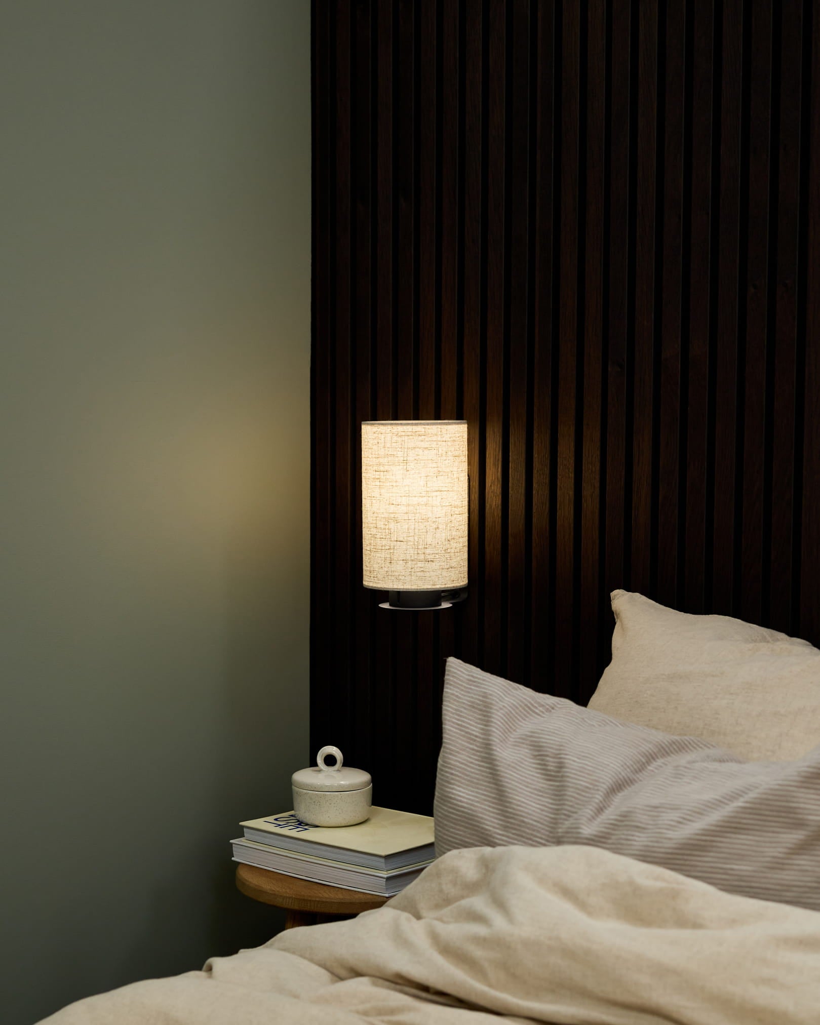 Rincón de dormitorio con un aplique de pared Feya de WoodUpp en Black/Beige, ropa de cama en beige y una mesita de noche con libros y una taza de cerámica con tapa.