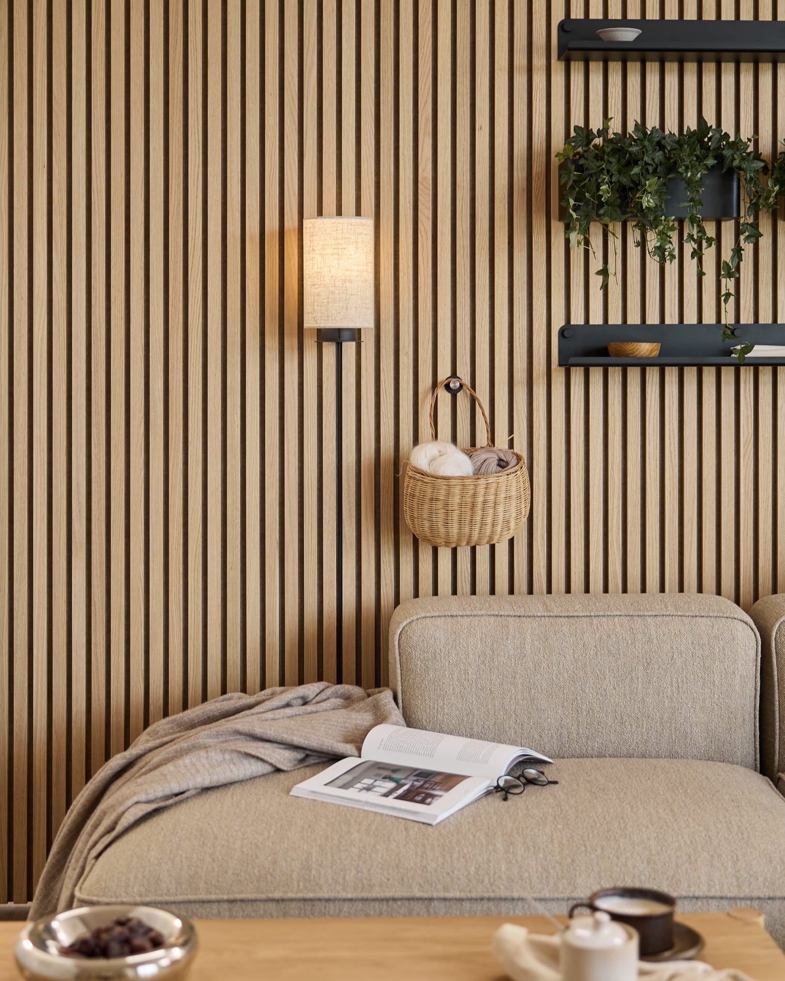 Sofá en Beige con manta y libro junto a una pared acústica de listones WoodUpp Akupanel, estantes con plantas y objetos decorativos, y un aplique de pared Feya en Black/Beige.