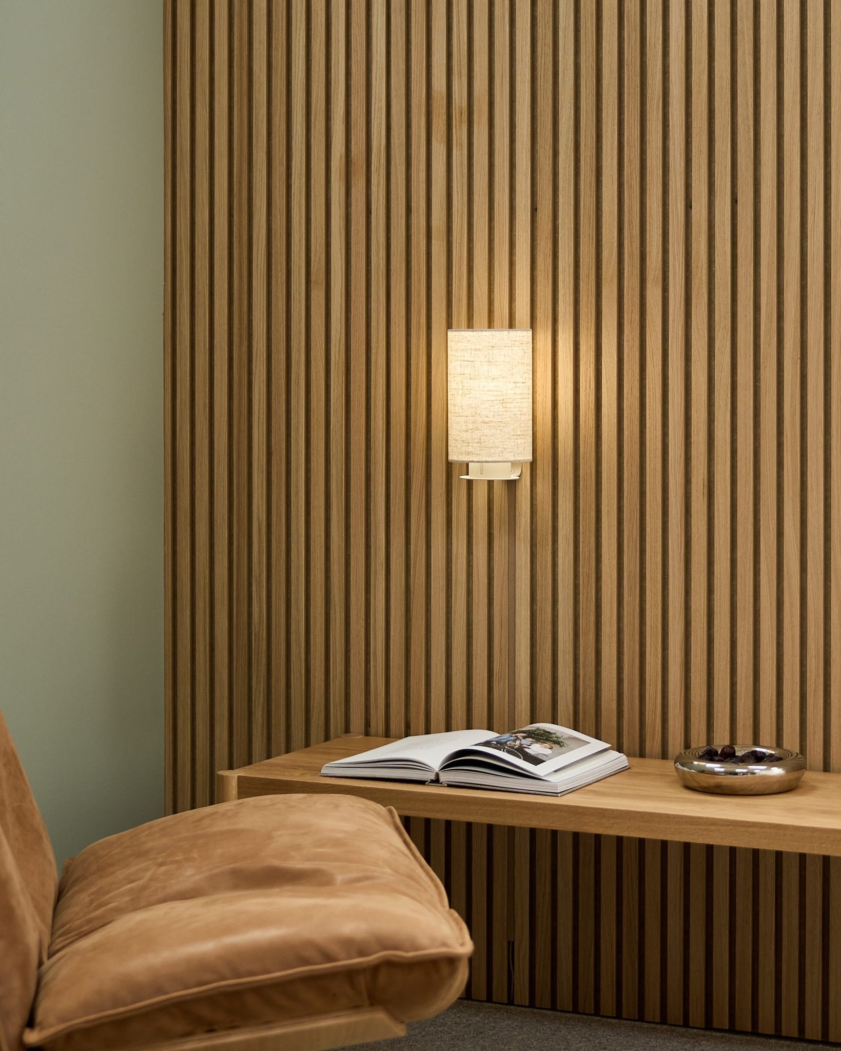 Espacio nórdico minimalista con una silla color canela, pared acústica de lamas WoodUpp Akupanel, lámpara de pared Feya en Beige/Beige, un libro abierto sobre un banco y un cuenco.
