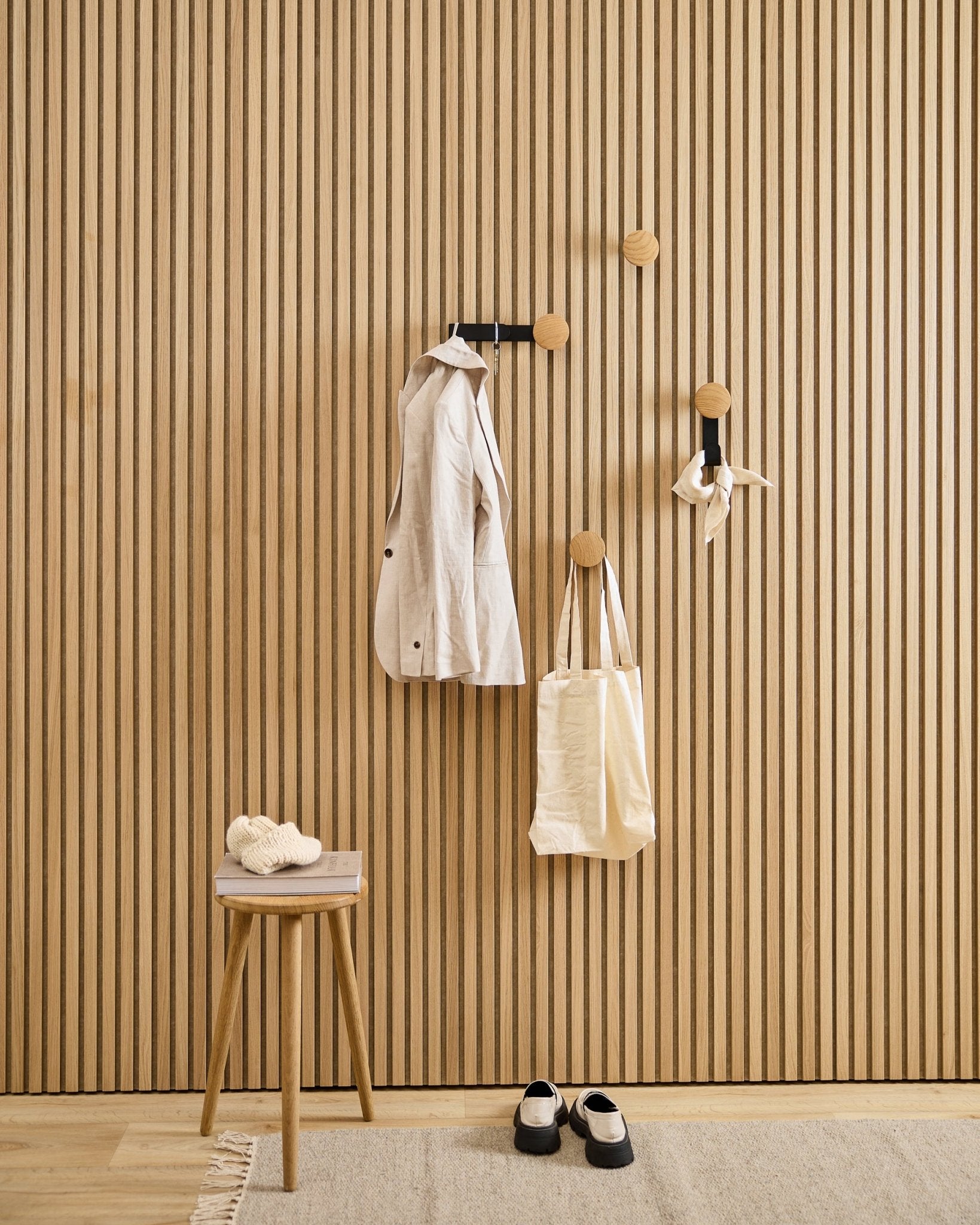 Recibidor minimalista con el colgador WoodUpp Linn Vertical en natural oak, con un abrigo beige, una bolsa tote, una bufanda, un taburete con zapatillas y zapatos black y blancos.