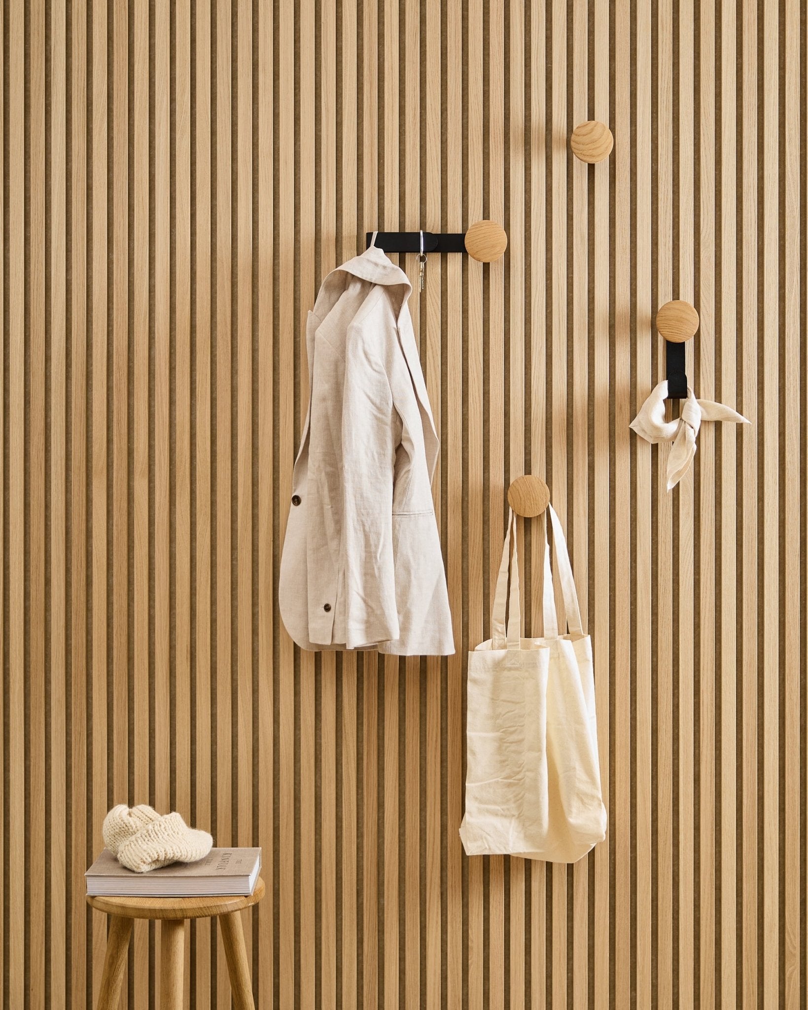 Una chaqueta de beige, un bolso tote y una bufanda cuelgan del Linn Horizontal Hanger de WoodUpp en Natural Oak; cerca, un taburete sostiene un libro y una prenda de punto.
