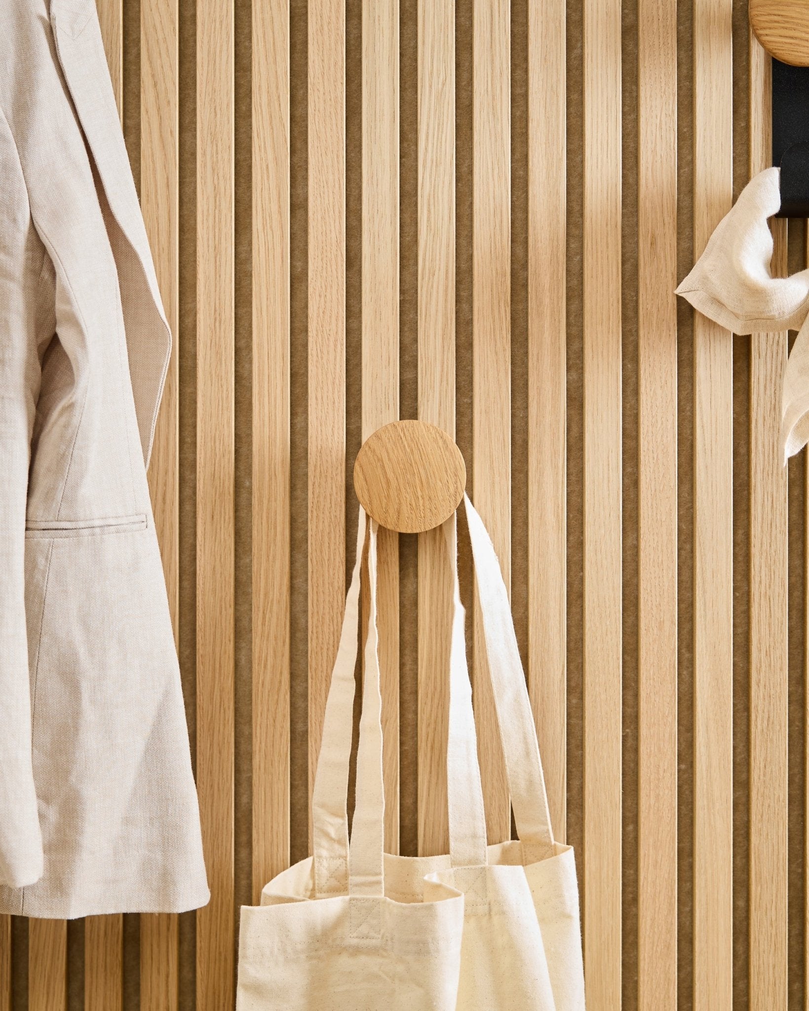 Un abrigo beige y un bolso tote cuelgan del Linn I Single Hanger en Natural Oak de WoodUpp, montado sobre un panel acústico de lamas de madera vertical Akupanel.