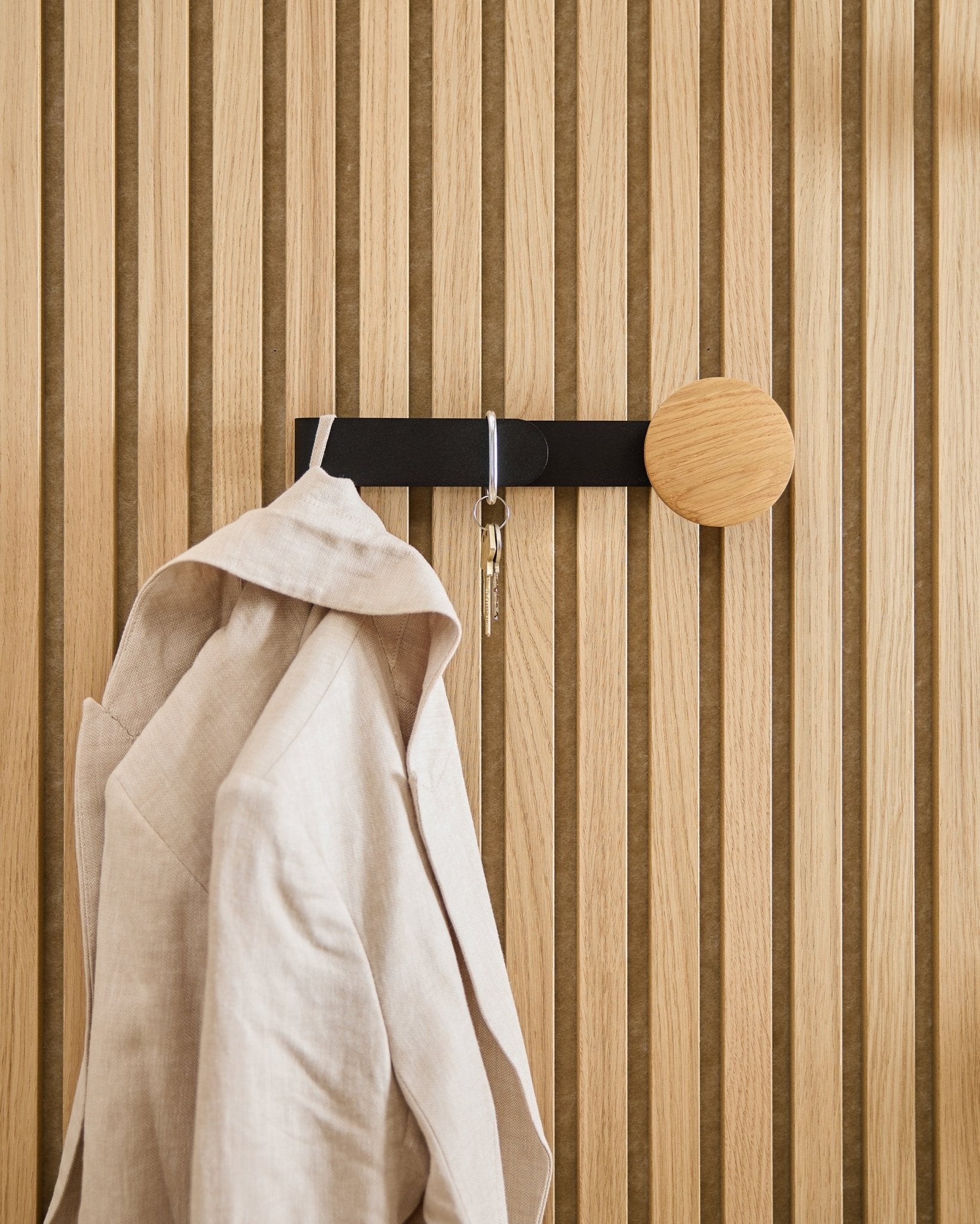 Una americana de beige y un juego de llaves cuelgan del Linn Horizontal Hanger de WoodUpp en natural oak, instalado sobre un panel acústico de madera Akupanel en natural oak.