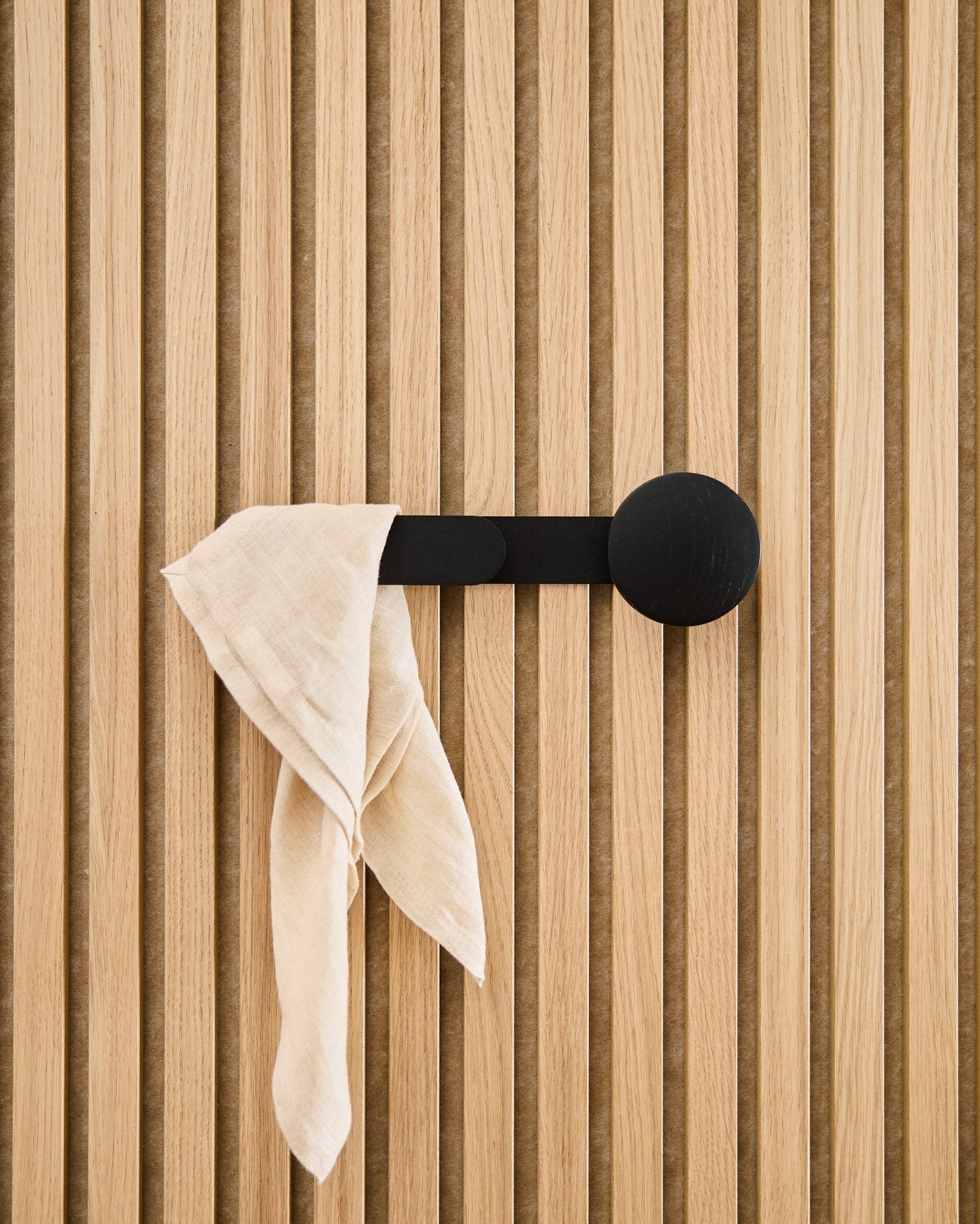 Una tela beige cae sobre el perchero WoodUpp Linn Horizontal en Black Oak, fijado a un panel acústico de listones vertical.